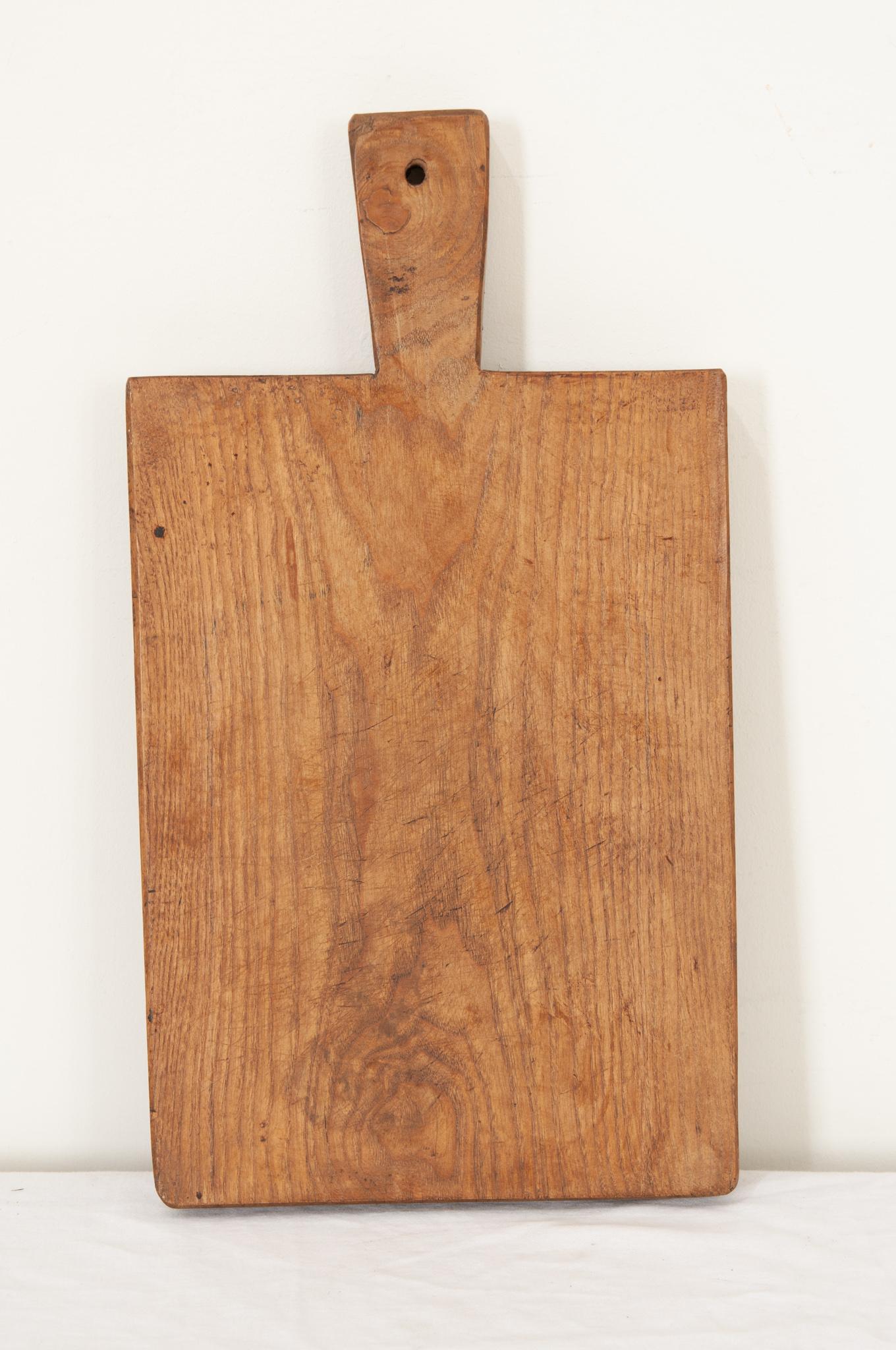 A solid wood cutting board from France in a rectangle shape with smooth corners and a handle. The worn board is made of a single piece of wood. Knife marks and scrapes that were left by cooks, now long gone, can be seen and felt as if they were made