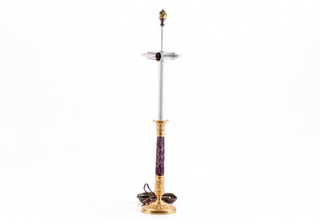 Lámpara candelabro dorada con motivos de hojas y liras de estilo neoclásico, y un fuste central de cristal tallado en tono amatista. Bombilla de araña doble con remate de llama dorada. 
Muy buen estado. 
Medidas de la lámpara: Diámetro de la base