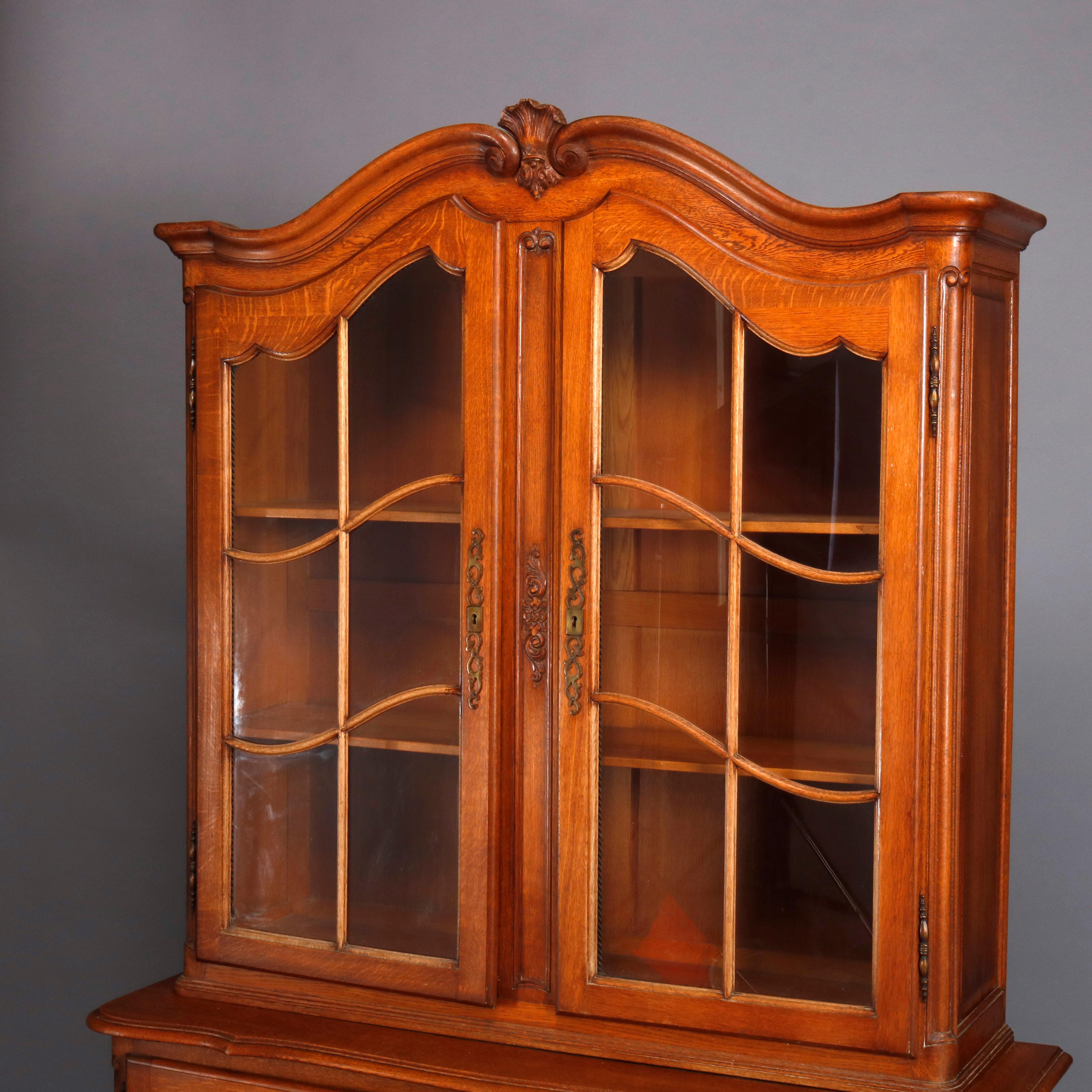 A French style breakfront cabinet offers oak construction with upper cabinet having shaped crest with central carved fleur-de-lis and flanking scrolled elements surmounting case with double glass doors opening to shelved interior, lower case with