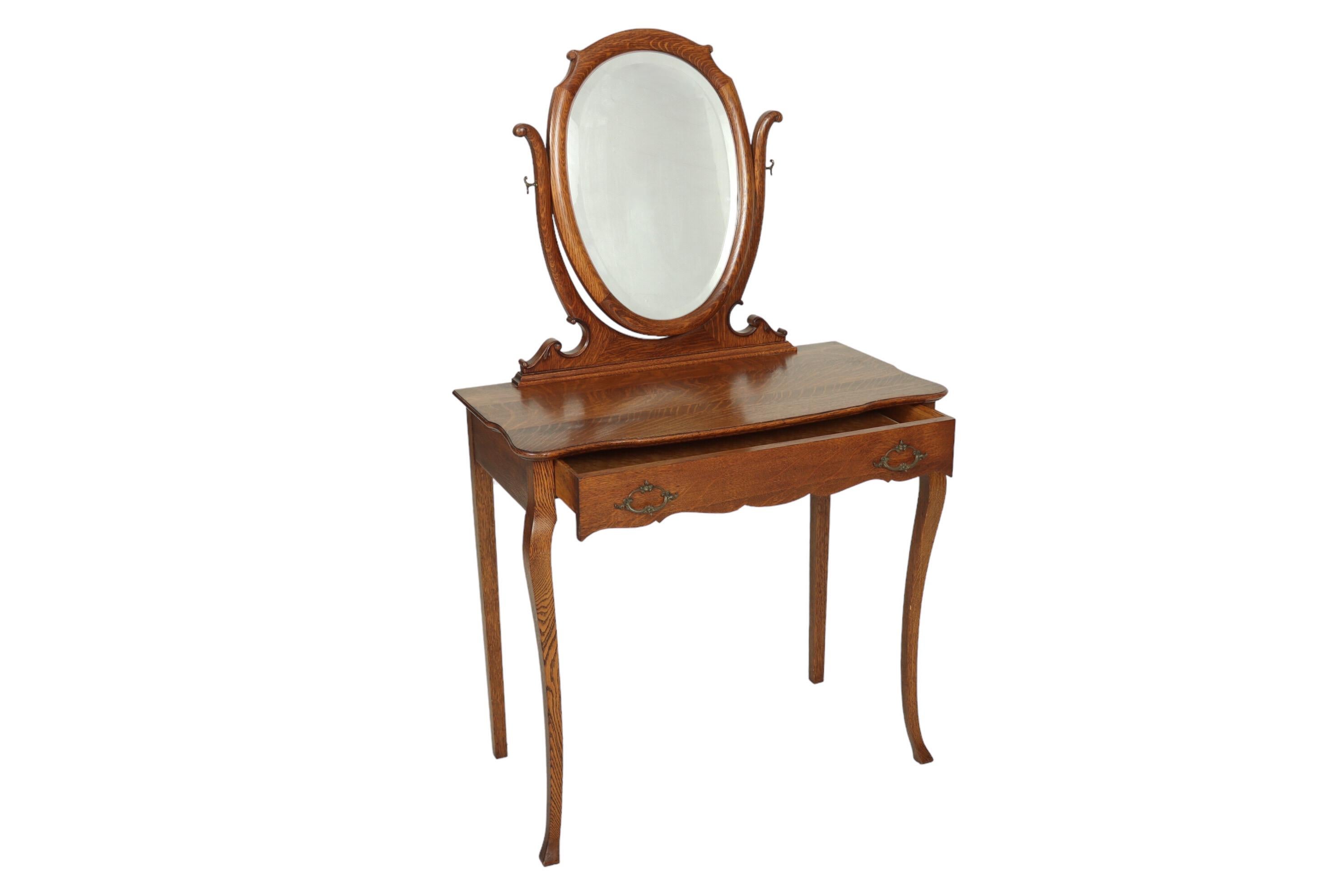 A turn of the century French style vanity made of tiger oak. An elegant oval beveled mirror is adjustable on swan neck supports. A single drawer with a serpentine edge opens with cast brass handles and is lined with birdseye maple veneer. Supported