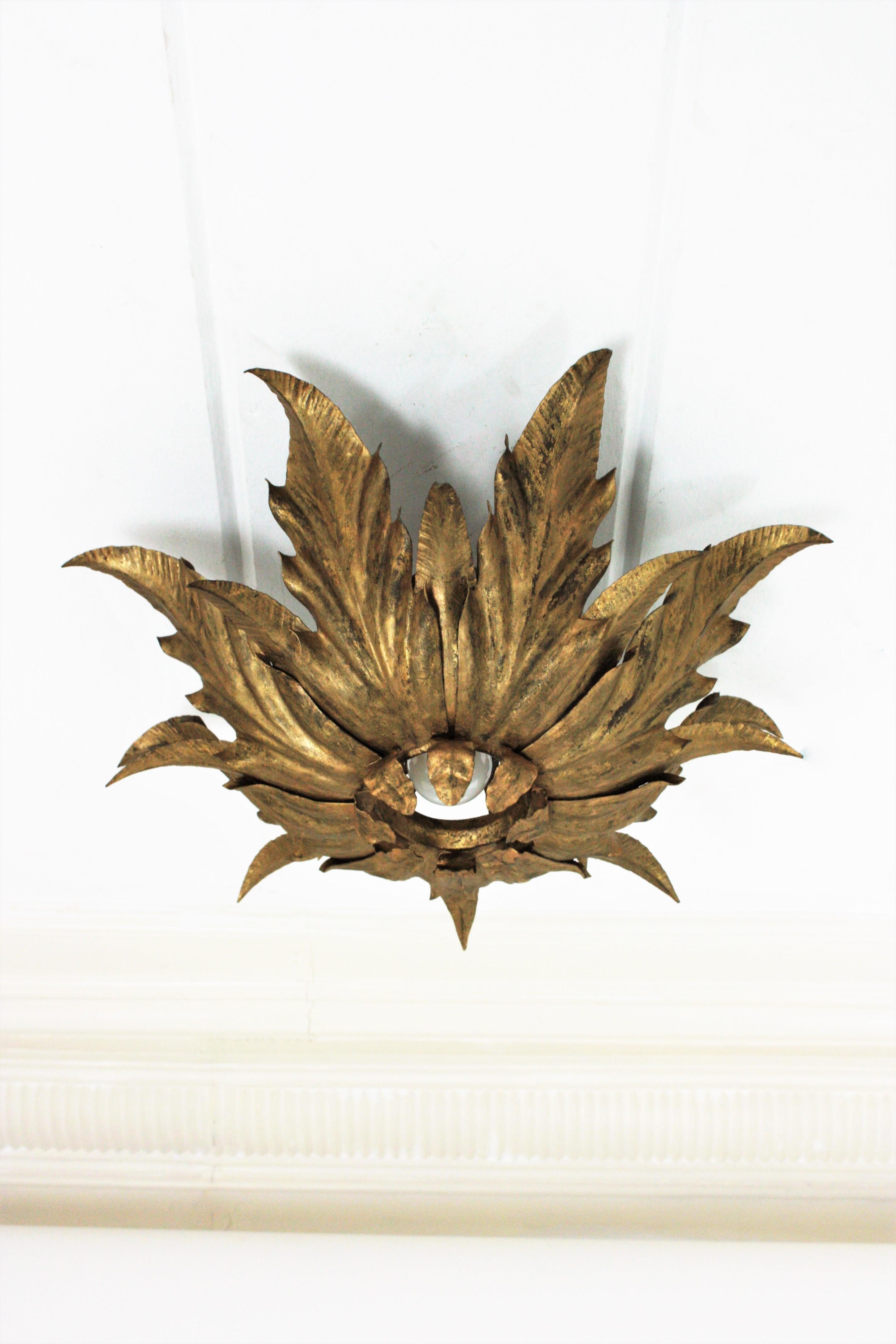 Stunning flower or sunburst ceiling light fixture, gilt iron, gold leaf, France, 1950s.
This eye-catching ceiling lamp features a double layered leafed frame of leaves surrounding a central exposed bulb. A layer of small curved leaves and a second