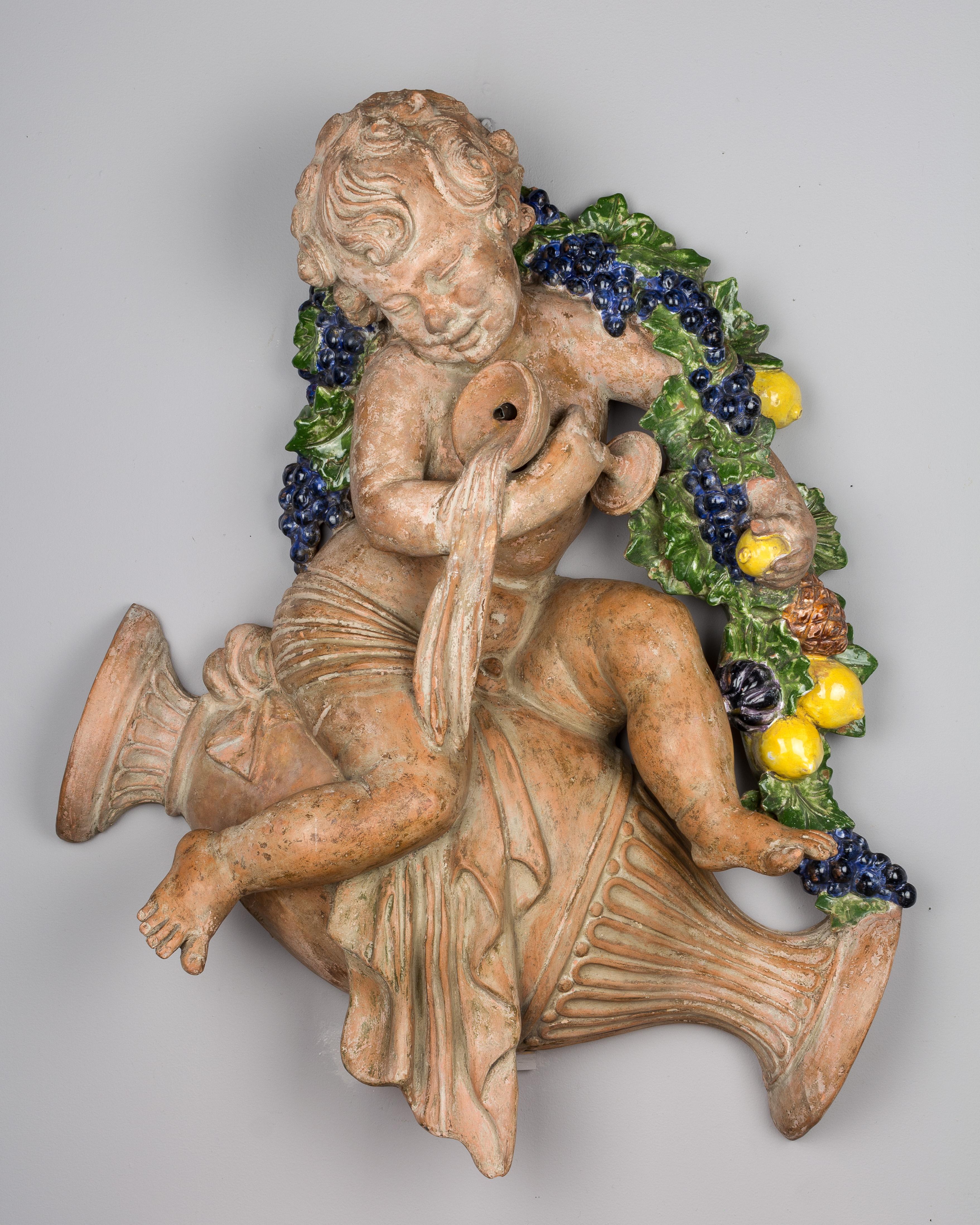 A French terracotta fountain in three parts, with a whimsical sculptural relief of a young Bacchus. Adorned with a colorful glazed terracotta garland of grapes and lemons, he sits astride a toppled urn, spilling his cup into the shell shaped basin