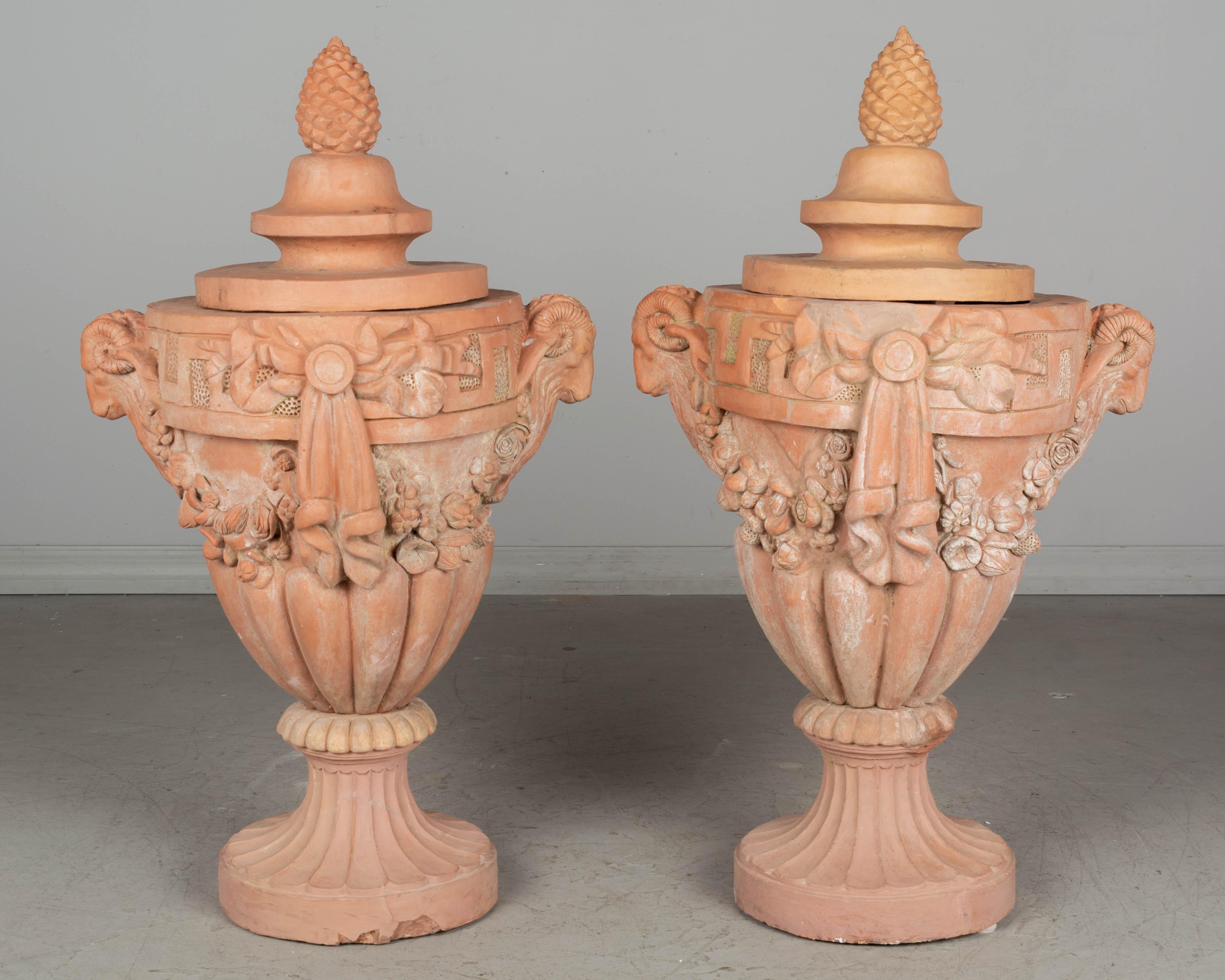 A pair of early 20th Century French Terracotta garden urns from the Toulouse Region. Nice casting with fine decorative details including large ram head handles, flower garlands with bows, and pinecone finials on the lids. Both urns have been