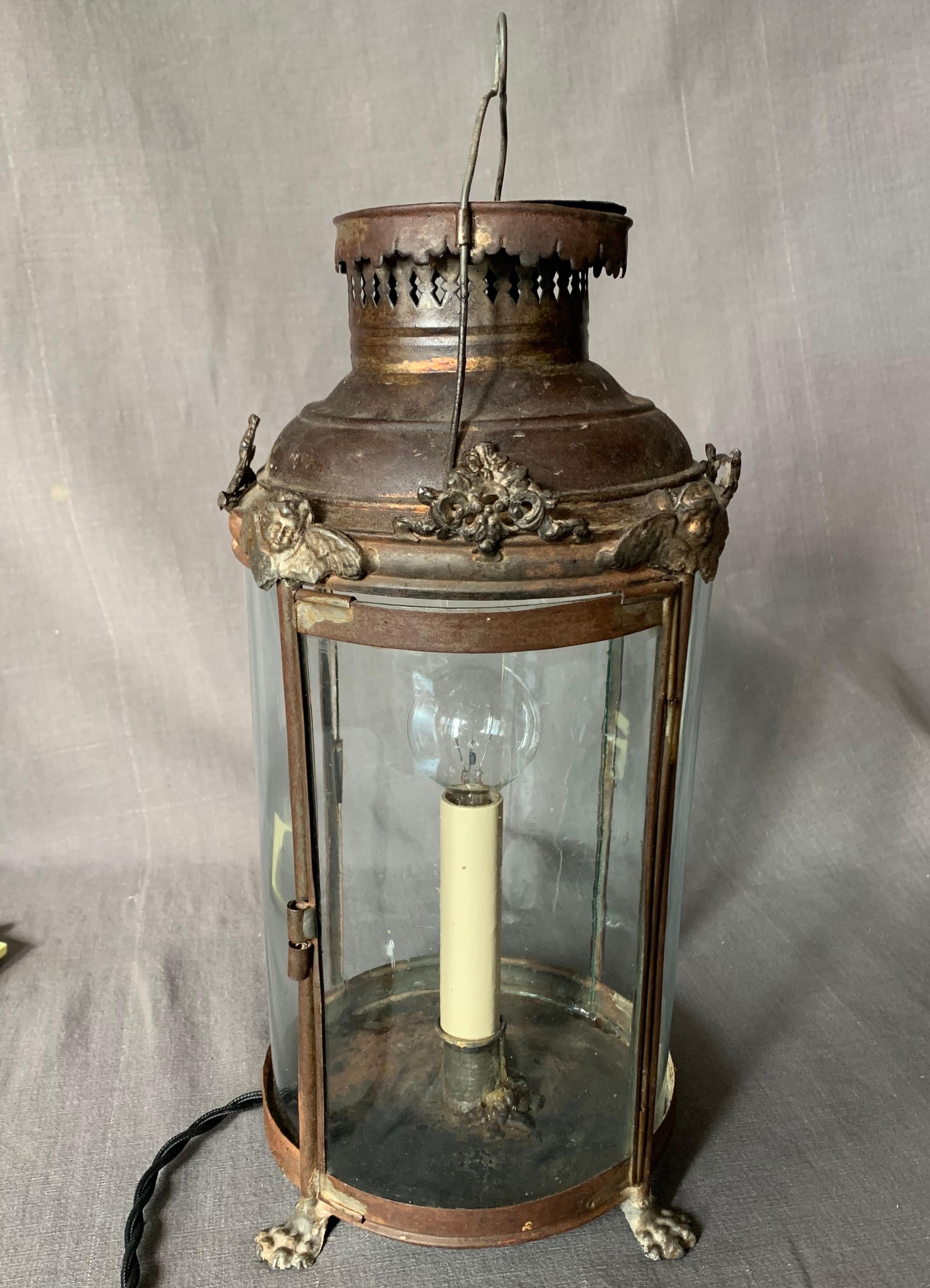 French tole lantern lamp. Antique brown patinated tole footed lantern candle stand with original early seeded curving glass panels with pierced ventilation cap surmounted with cherubs and foliate scrollwork, newly electrified with twisted silk cord