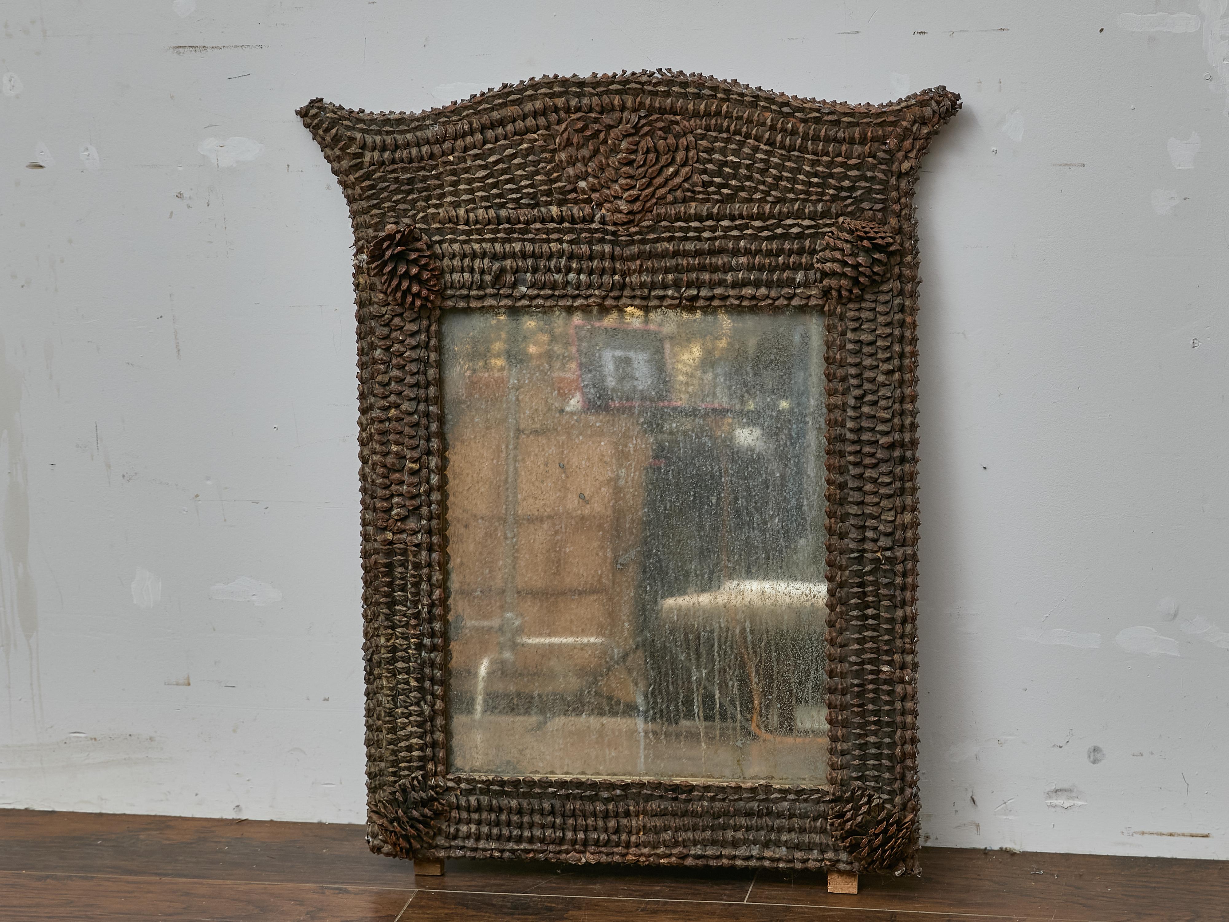 Questa espressiva specchiera francese, risalente al 1900-1920 circa, presenta un'impressionante cornice Tramp Art intricatamente decorata con scaglie di pigna. Lo stemma ad arco e gli angoli scultorei conferiscono allo specchio un'inconfondibile