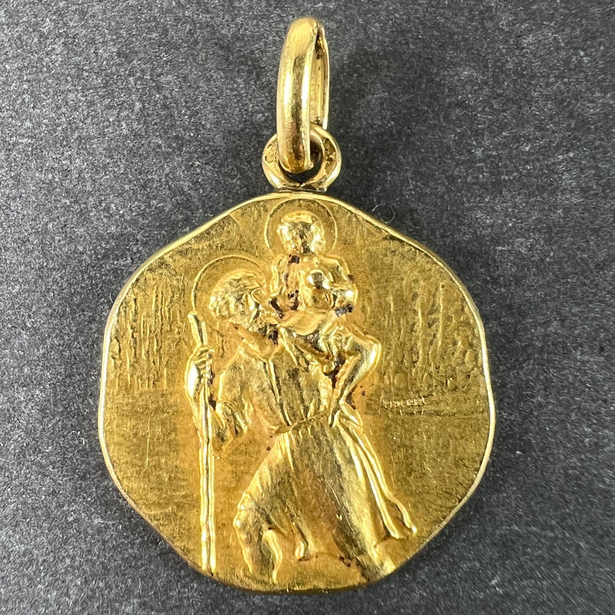 An unusual French 18 karat (18K) yellow gold charm pendant designed as a medal depicting Saint Christopher as he carries the infant Christ across a river with a forest and mountain scene on the other side, including a small house by the bank of the