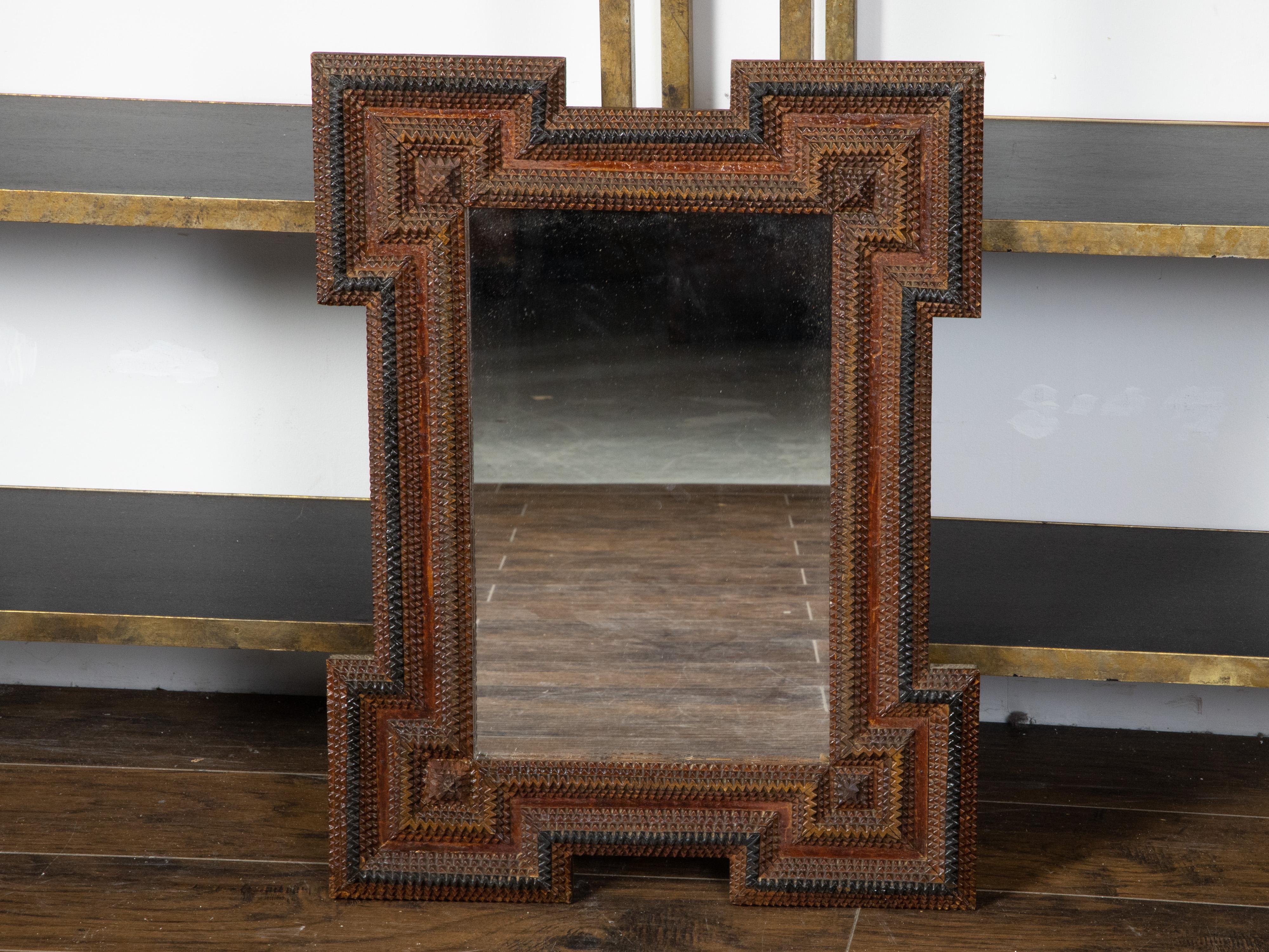 A French Tramp Art hand-carved wooden mirror from the early 20th century, with raised motifs, dark patina and protruding corners. Created in France during the turn of the century, this French Tramp Art mirror features a linear frame with raised