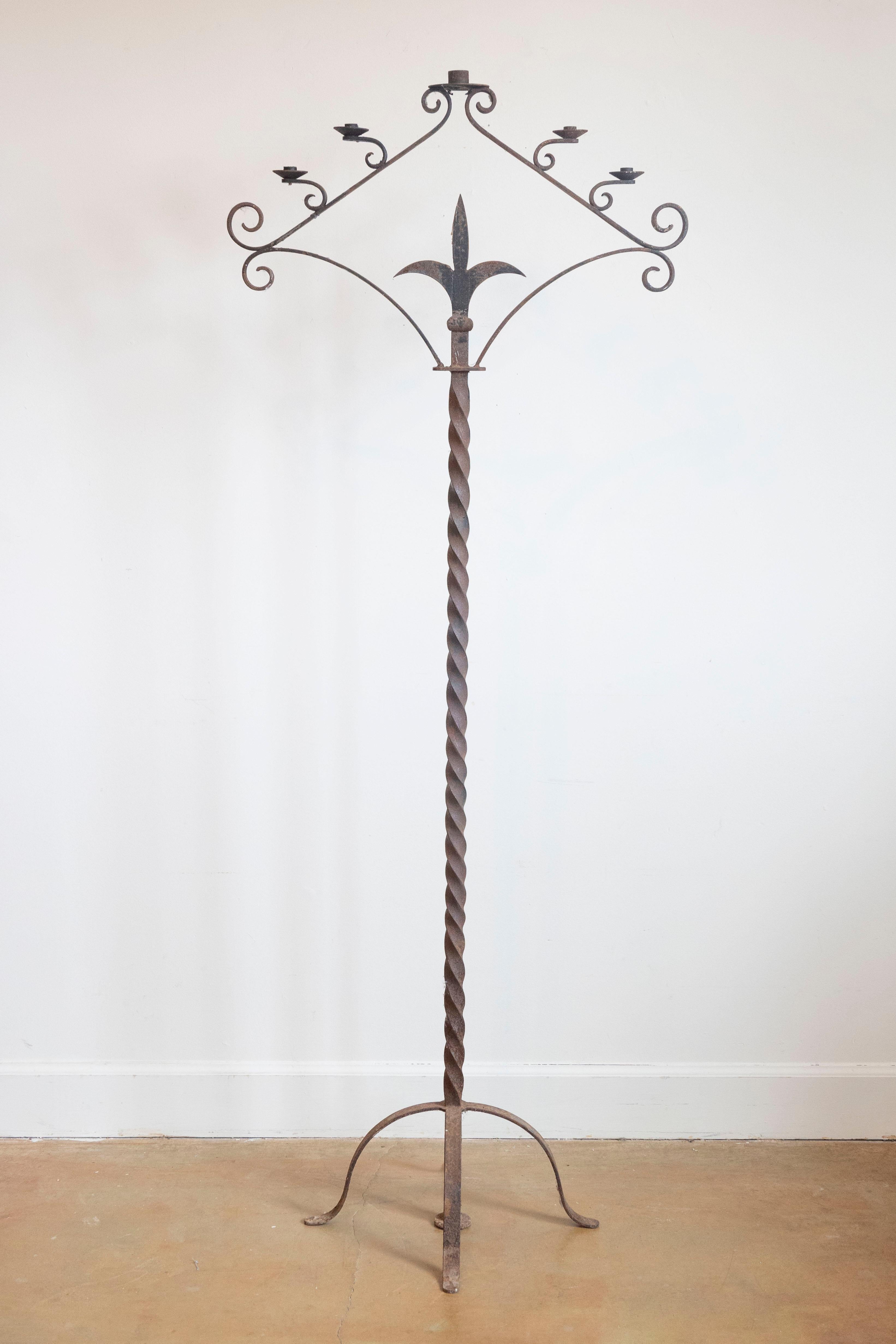 Un alto candelabro da terra francese in ferro dei primi del Novecento, con cinque pinnacoli, disegno a volute, motivo stilizzato a fleur de lys e accenti ritorti su una base quadripartita. Creato in Francia a cavallo tra il XIX e il XX secolo,