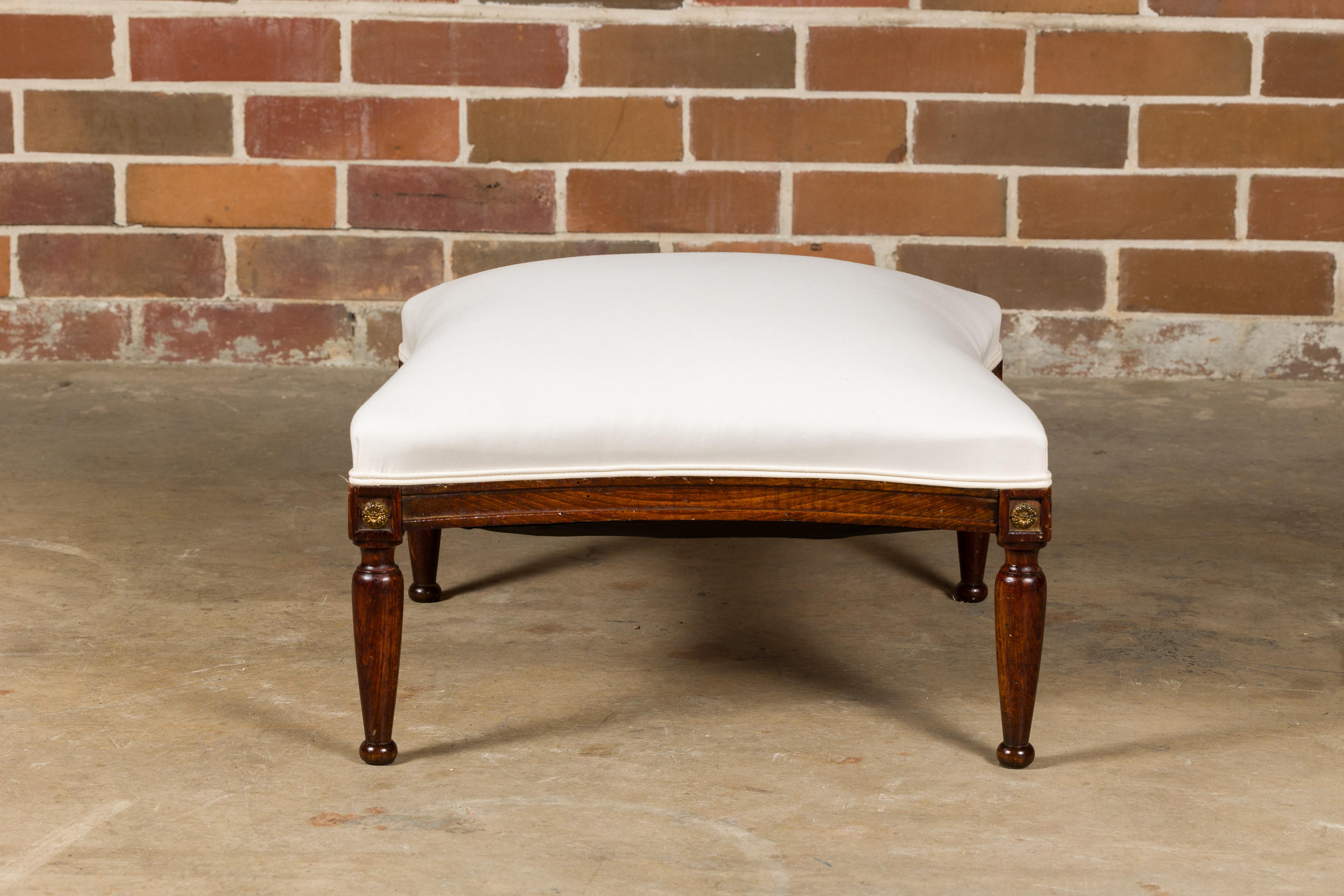 A French wooden ottoman from circa 1900 with tapering cylindrical legs, carved giltwood rosettes and muslin double welt upholstery. Step into the allure of French sophistication with this charming wooden ottoman from circa 1900, a piece that marries