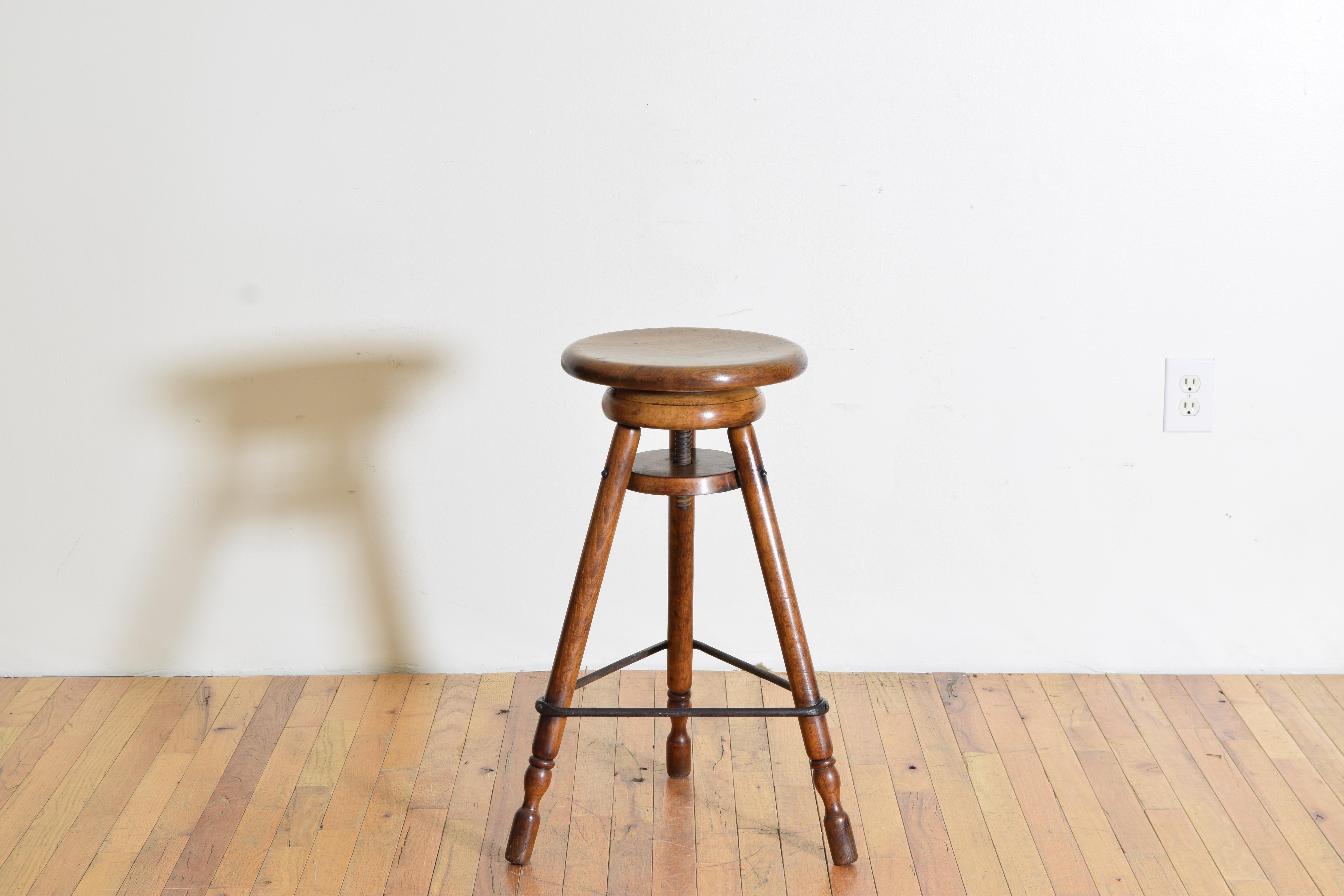 Constructed entirely of solid walnut and having a rotating slightly concave circular seat atop an adjustable down rod supported by three legs braced in iron and ending on uniquely shaped feet, height measured to lowest possible setting, diameter is
