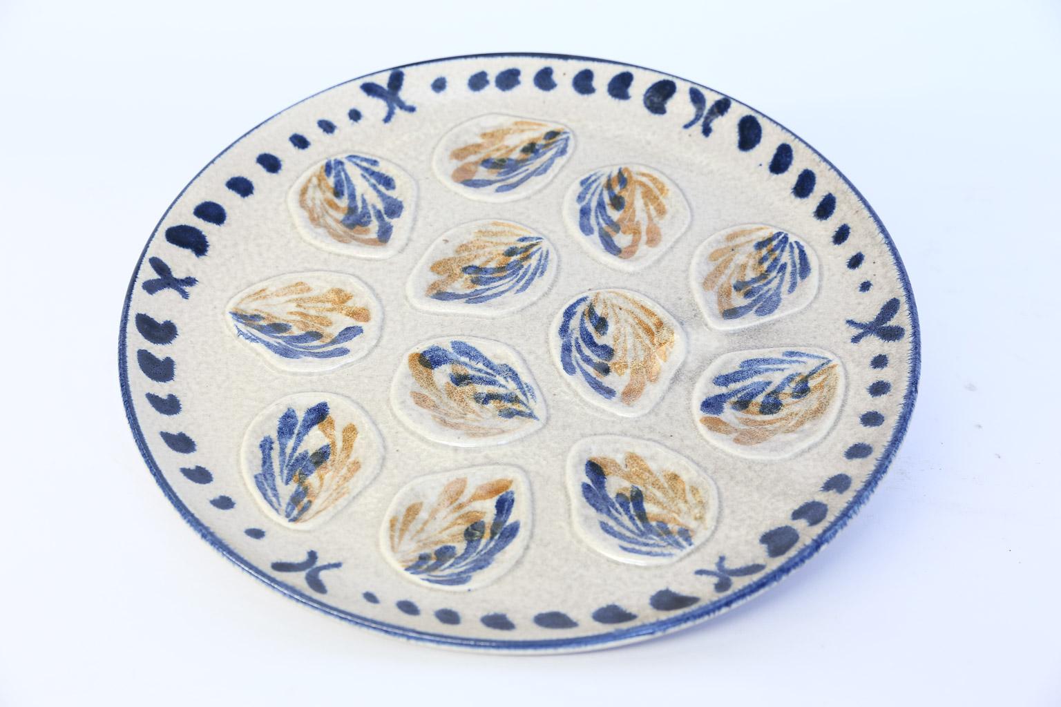 This is a wonderful French twelve well oyster platter. The platter has twelve wells for serving oysters. The hand painted pattern depicts leaves in blue and orange on a beige background. Although there is no mark on the back, this platter was made