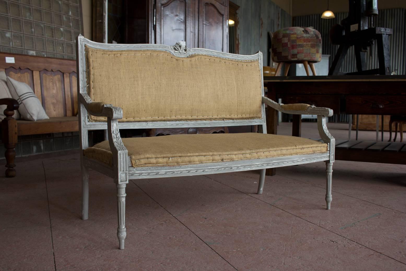 Antique French Louis XVI style two-seat settee the carved ribbon cresting top rail above a hessian upholstered padded back, seat and arms raised up on turned fluted legs.