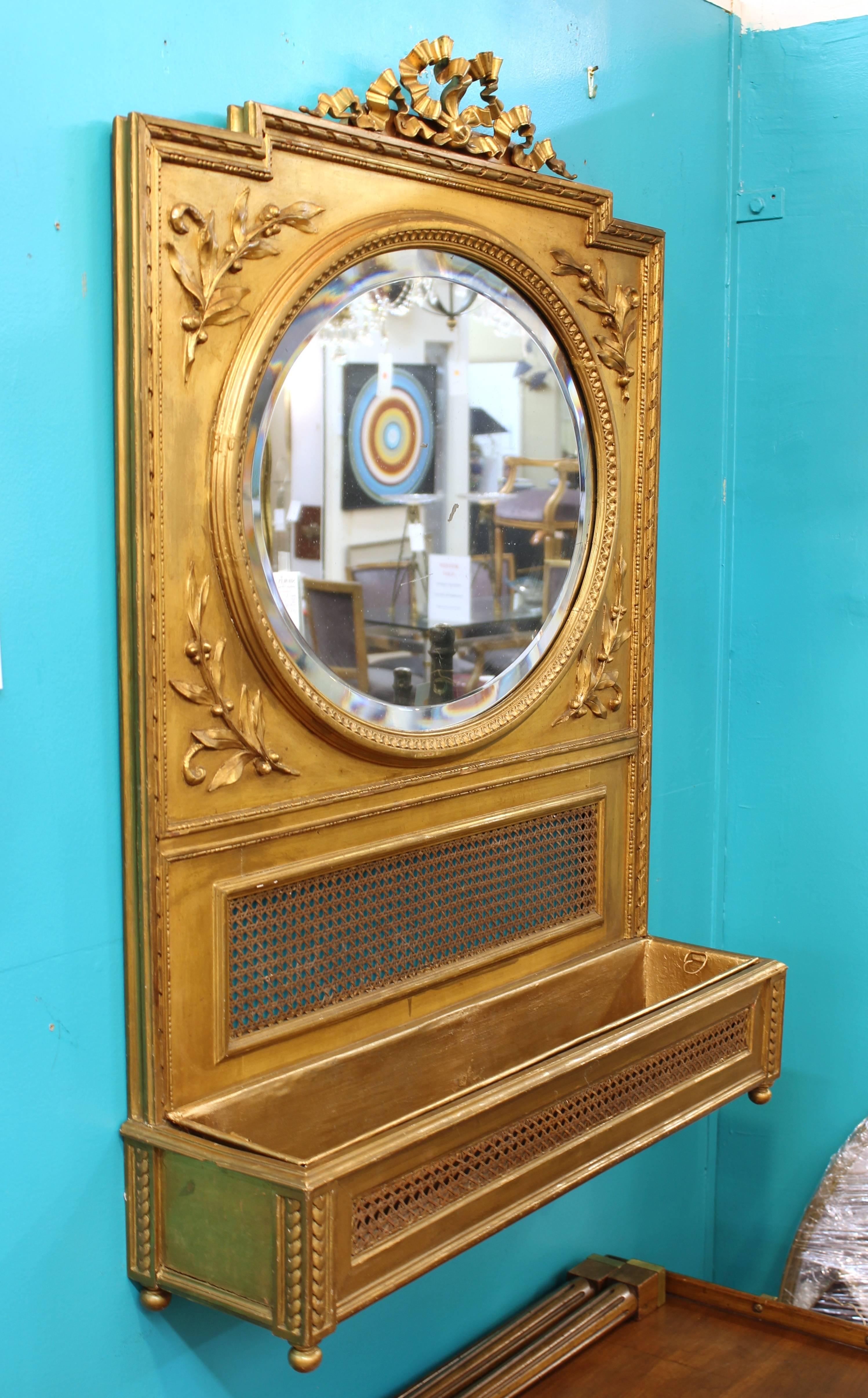 A Victorian gilded round bevelled mirror with planter, in neoclassical style. The wood is hand-carved and caned, and has a bronze fragment on top that is reminiscent of the style of Louis XVI.
The piece was made in France in the 1880s and is in