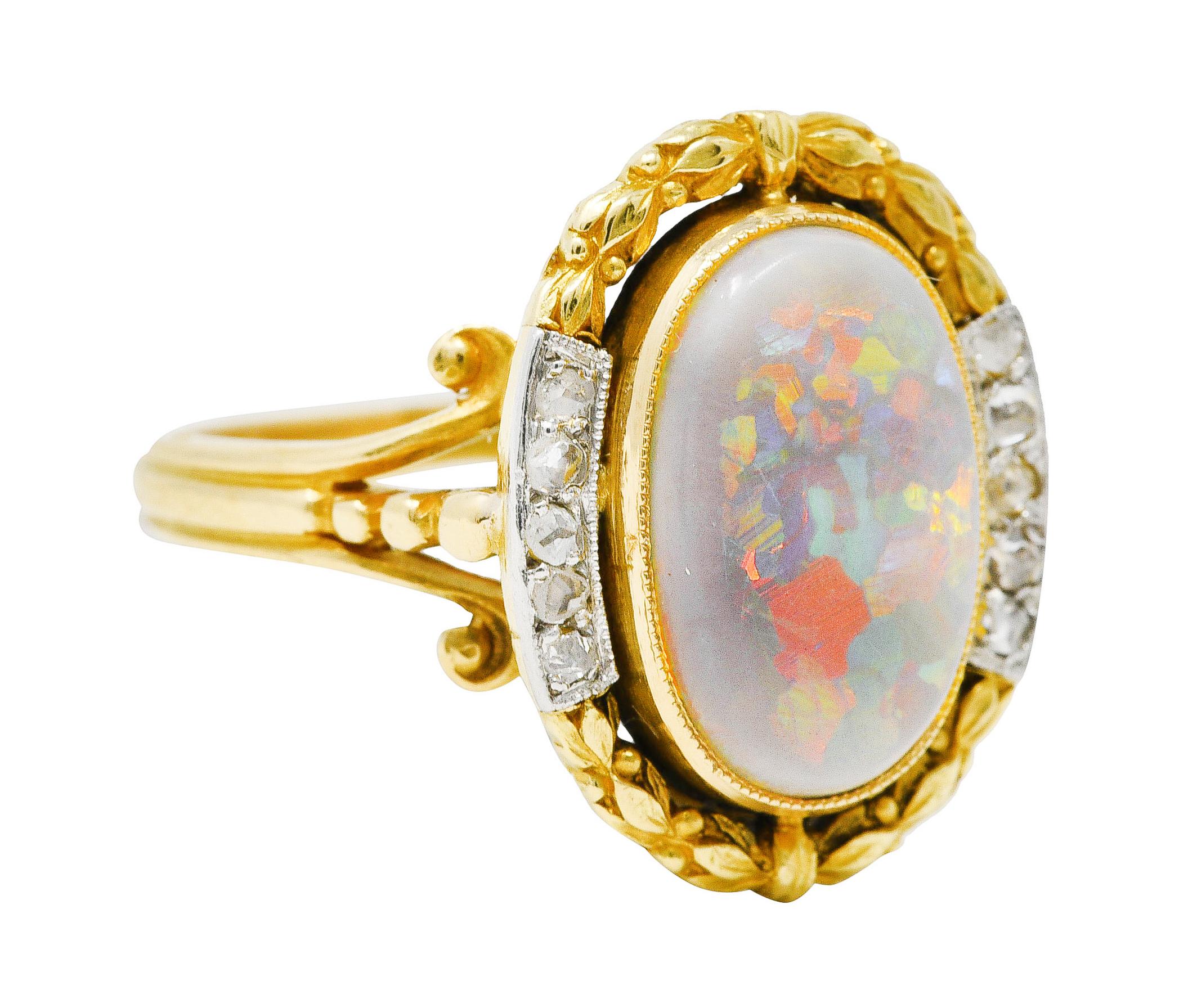 Centering an oval opal cabochon measuring 8.0 x 13.0 mm - gray in body color with spectral play-of-color. Bezel set with a highly rendered gold laurel motif surround with platinum-topped sections. With bead set rose cut diamonds weighing