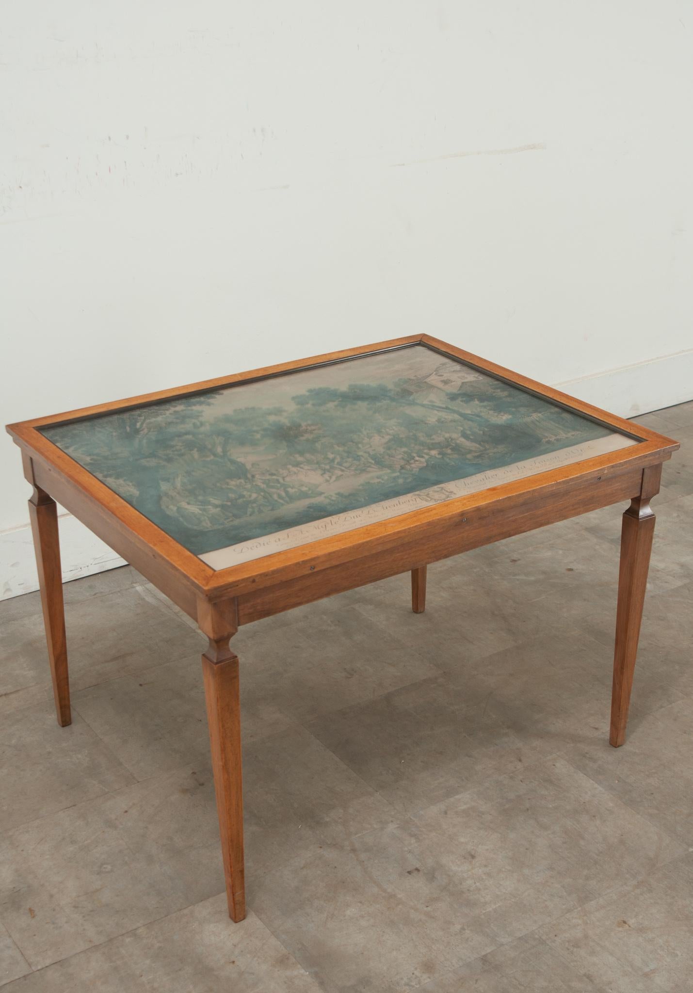 A vintage coffee table with a mahogany frame and glass top. This vintage table features a French lithograph underneath the glass that can be changed out to display the art of your choice. The top is raised on four tapered legs. Cleaned and polished