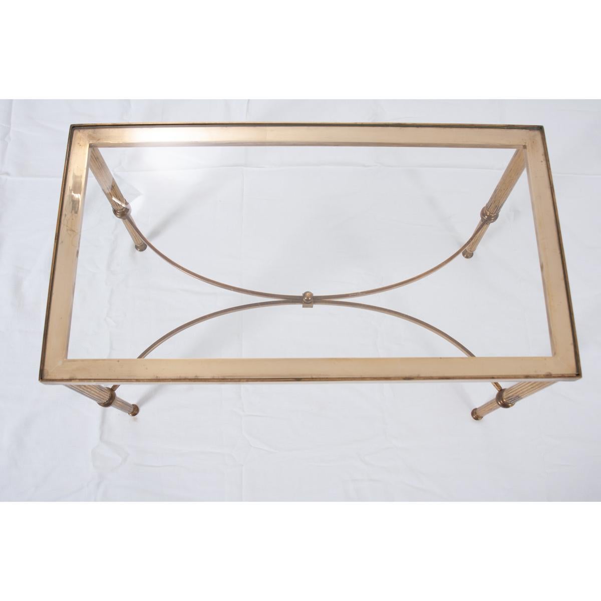 This stylish brass cocktail table or coffee table boasts a clean design that is simply stunning. The delicate brass stretchers form a soft, sweeping X with a brass finial in the center. The thin brass legs are reeded with trumpet-like feet.