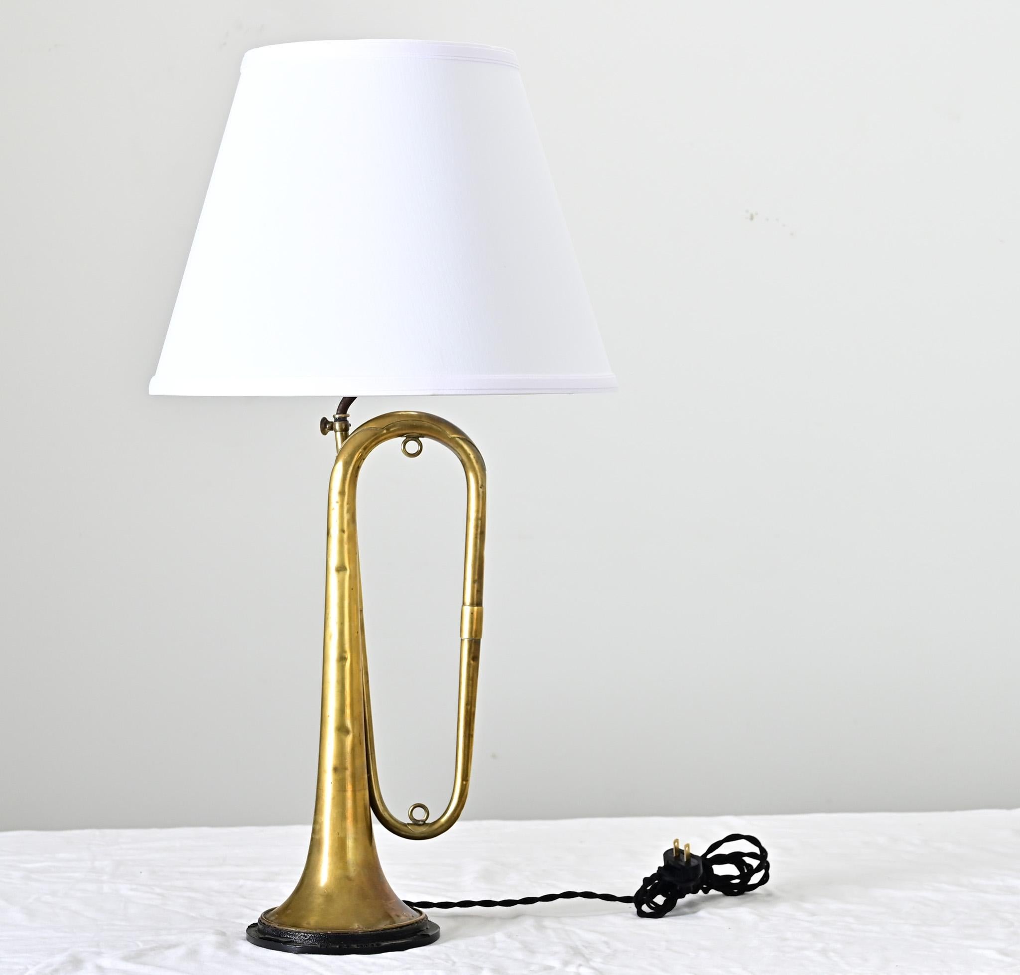 Una trompeta de latón Couesnon vintage fabricada en Francia que se ha reciclado para convertirla en una lámpara de mesa única. La trompa muestra signos de haber sido un instrumento musical muy querido y utilizado; el latón está oscurecido y abollado