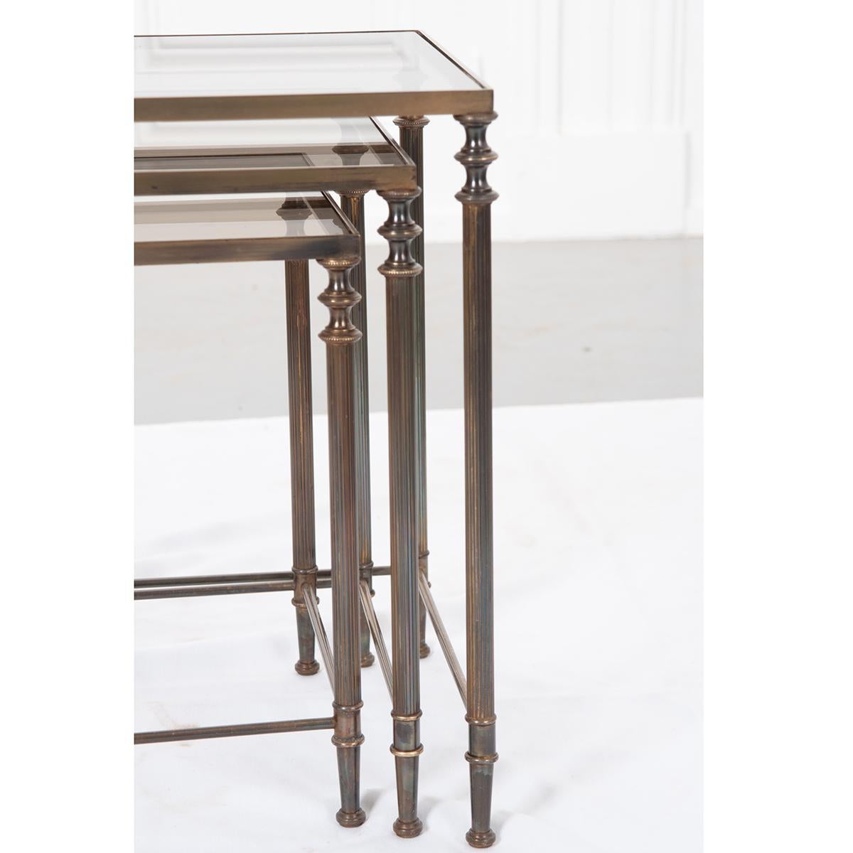 Set of three French, brass nesting tables with inset tinted glass tops. The four legs have turned-like caps with fluted columns and turned-like feet. Simple brass rod stretchers connect the legs for an added detail. 

Dimensions:
1. 17-¼”H x 20-⅞”W