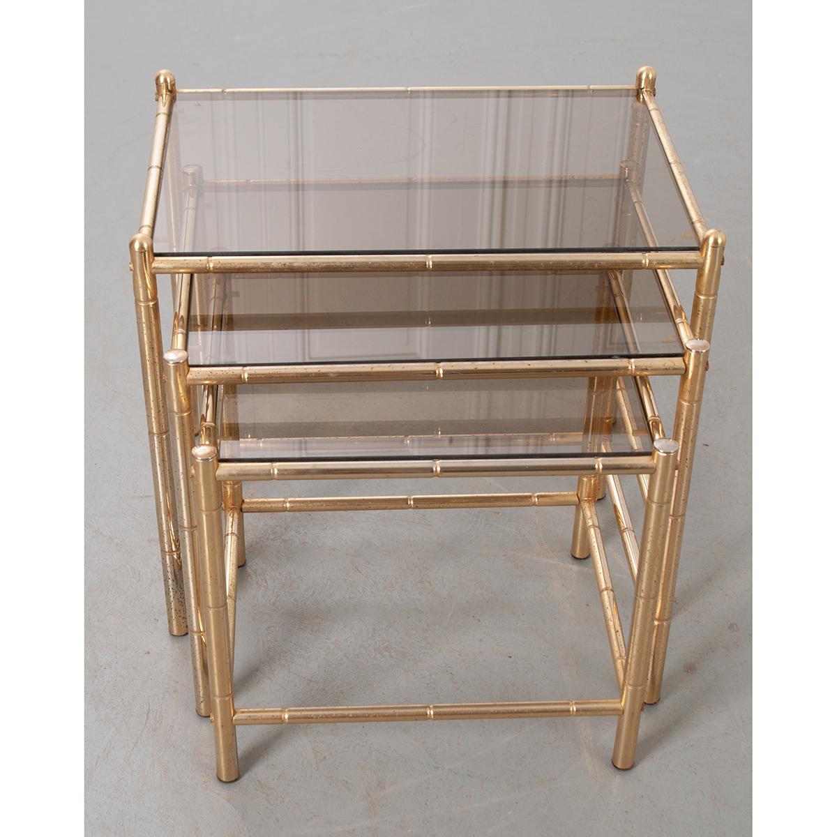 Set of three French, brass nesting tables with inset, tinted glass tops. The brass plated frames are styled to mimic bamboo and have a lustrous metallic finish. The tables conveniently nestle into one another, minimizing their footprint when not in