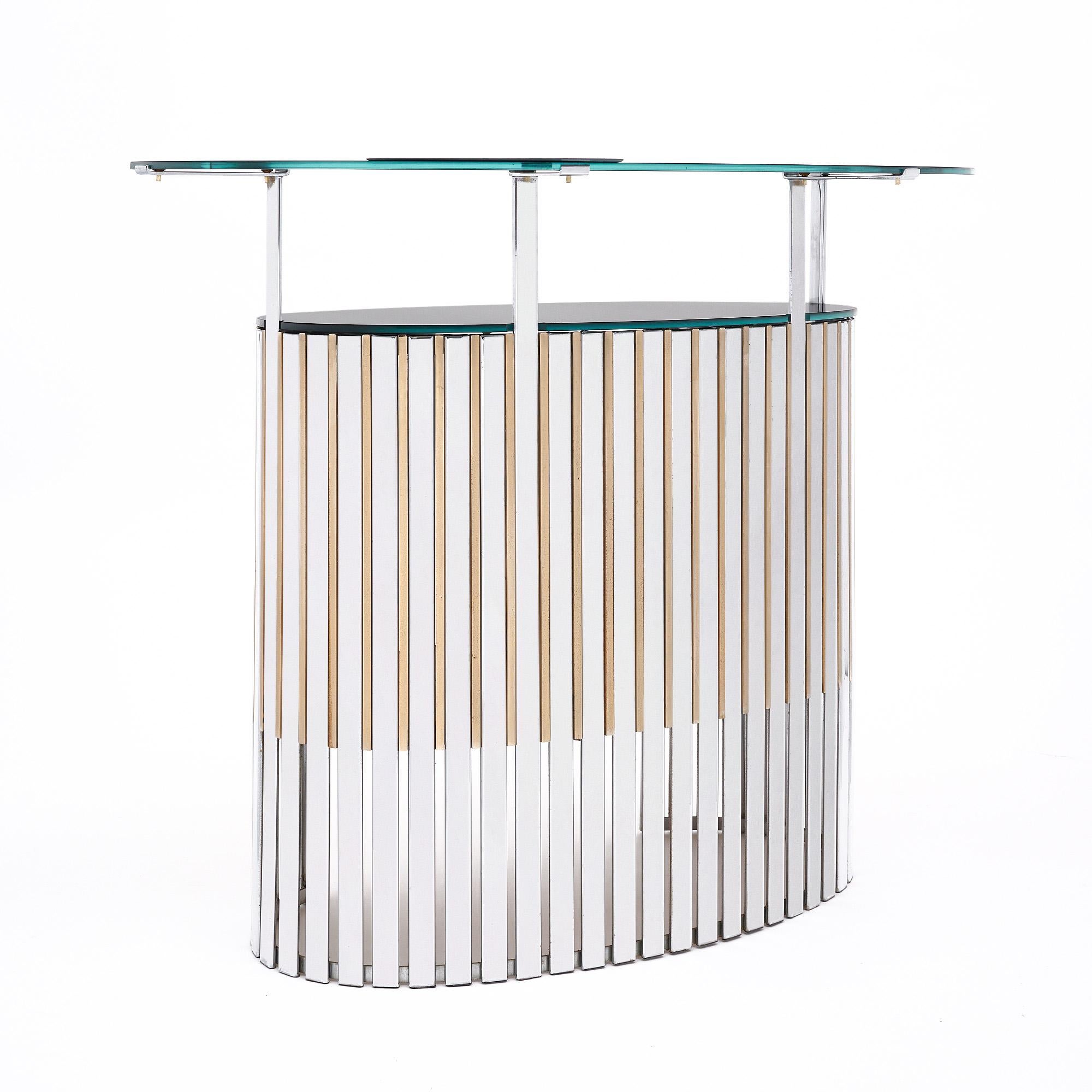 French cocktail bar in an oval shape, crafted with alternated blades made of chromed steel and solid brass. The bar is finished with a graphite mirrored top and an arched counter. The bar features a mid height inside shelf.
