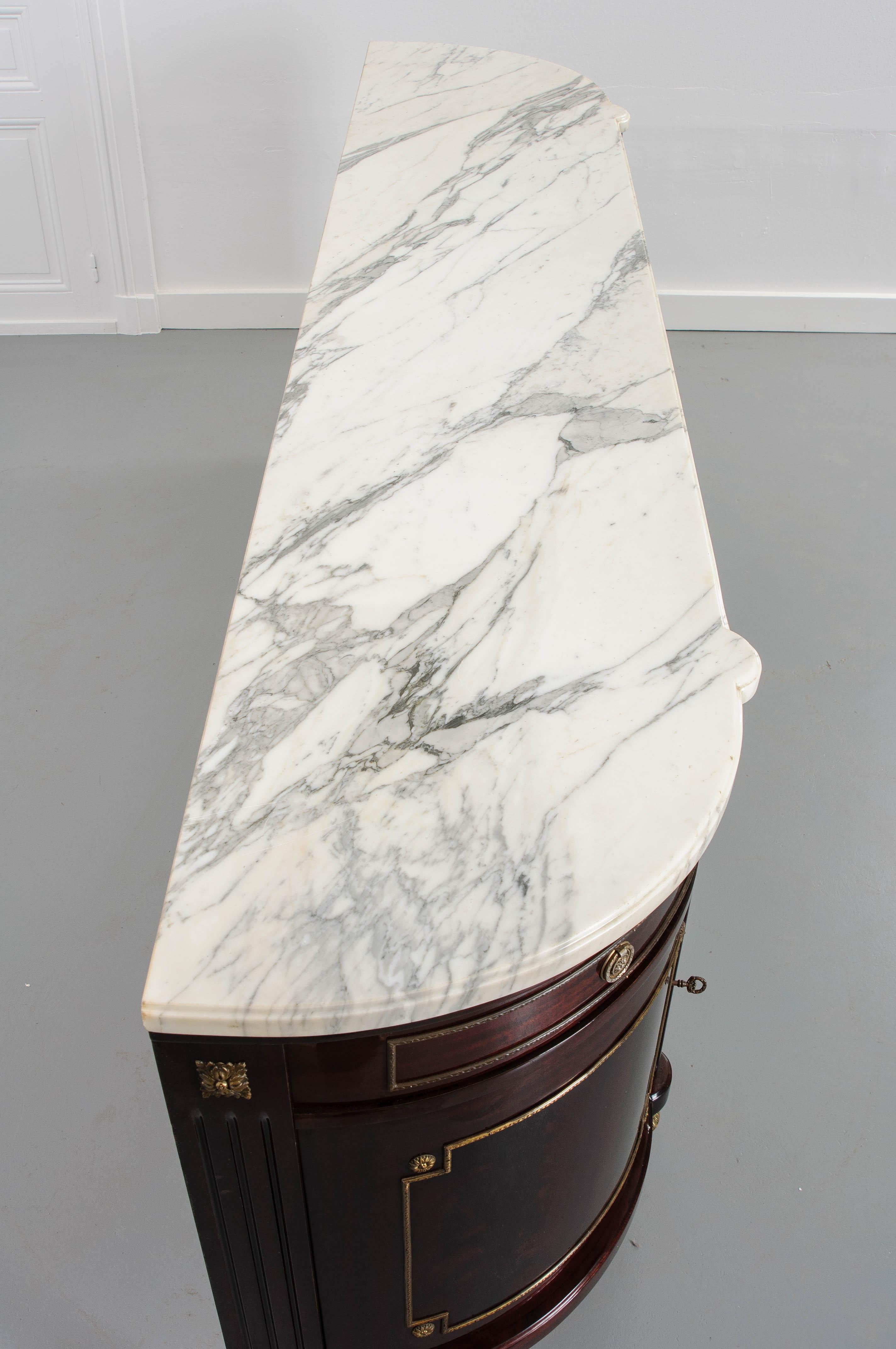 This fine Louis XVI-style vintage marble-top demilune enfilade, circa 1950s, features a beautiful high-gloss finish which highlights the exquisite bookmatched mahogany. The carved white marble top with grey veining rests above an apron of five short