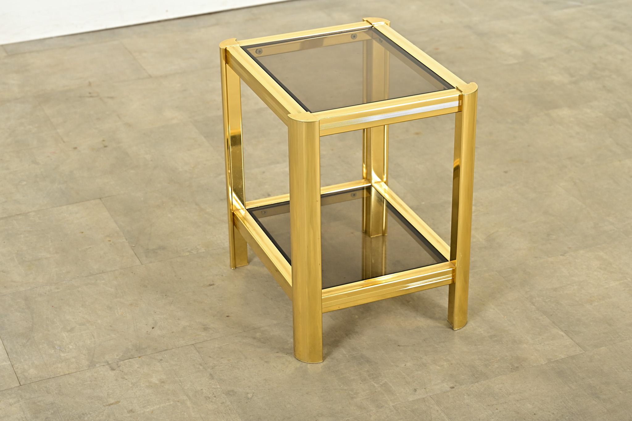 Fabulosa mesa auxiliar o auxiliar de dos niveles de los años 70, con estructura de latón pulido e inserciones de cristal ahumado. Una franja de junquillos acentúa las líneas rectas de la parte superior y los laterales, mientras que las patas de