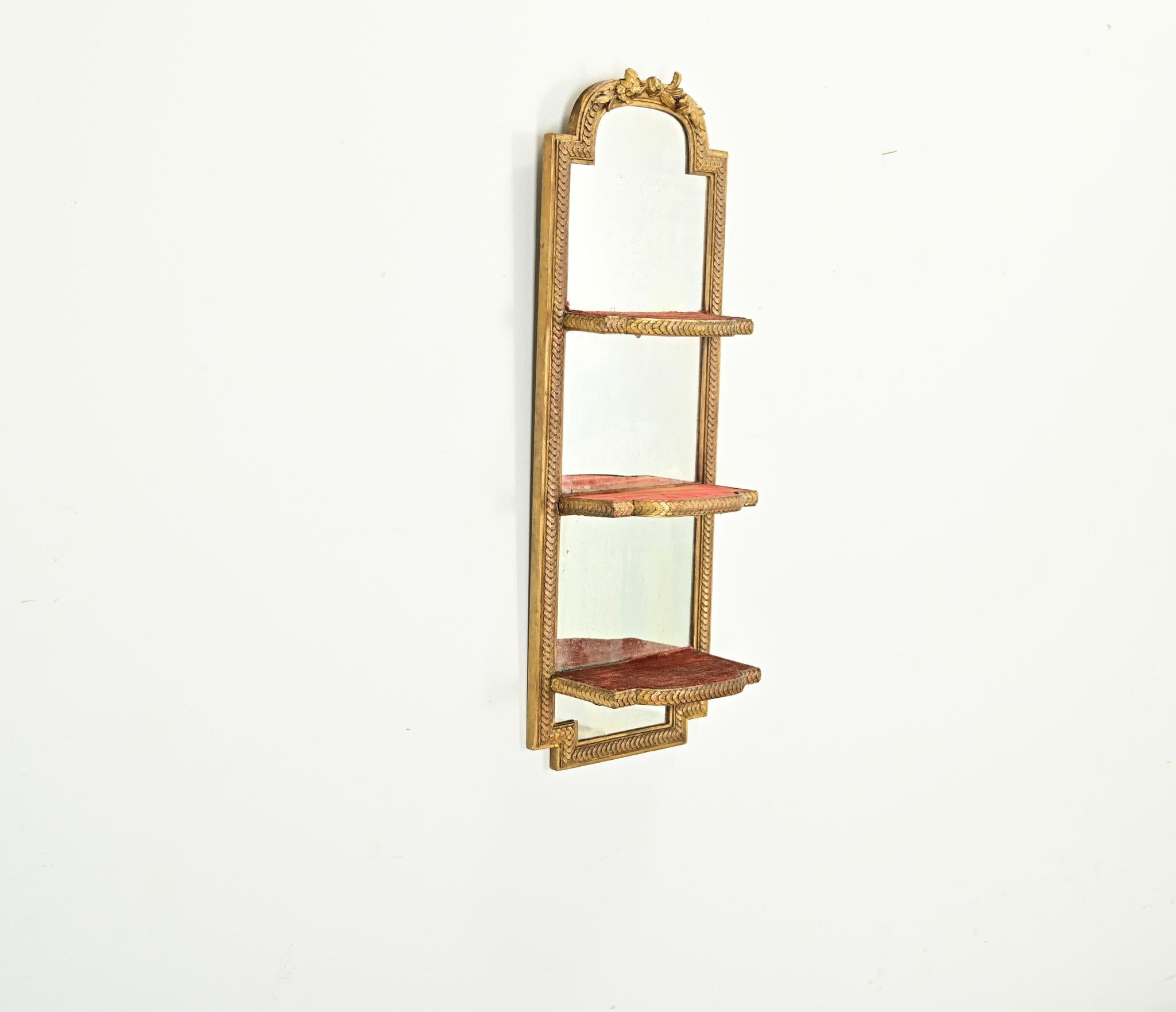 French vintage 20th century mirror-back hanging wall shelf in Louis XVI style gold gilt frame. The arched top of the mirror is crowned with a floral garland, transitioning into a layered, fishscale pattern that wraps the molding and each shelf. The