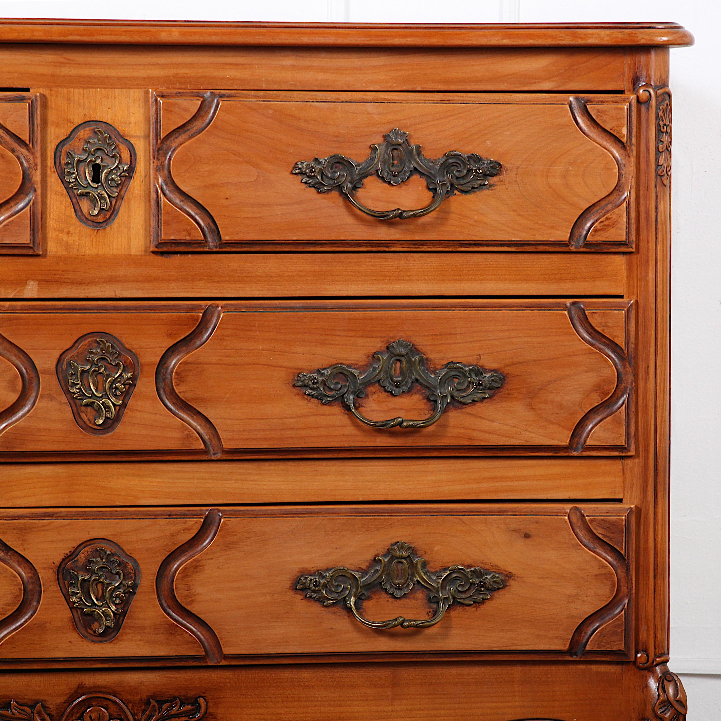 A mid-20th century French vintage Provencal solid cherry wood commode with two short drawers over two long, the sides with shaped panels and the bow-front with carved details to the drawers and the original brass hardware. Raised on elegant scrolled