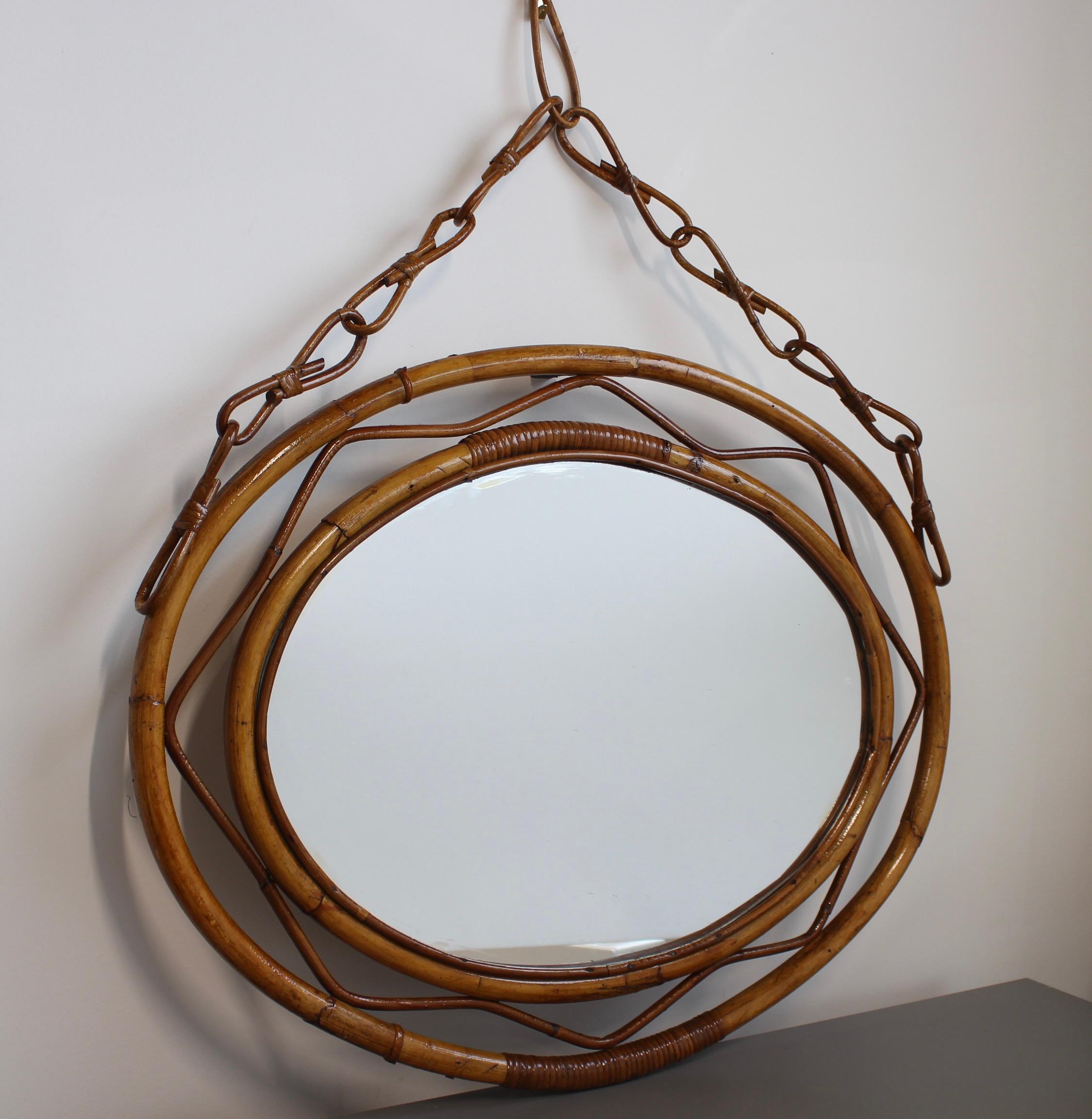 Miroir mural vintage en rotin de forme ovale (circa 1960). Ce miroir a un design très charmeur avec deux cercles concentriques en rotin encadrant le miroir. La chaîne de suspension ajoute de l'allure et de la hauteur à l'ensemble. Le cadre présente