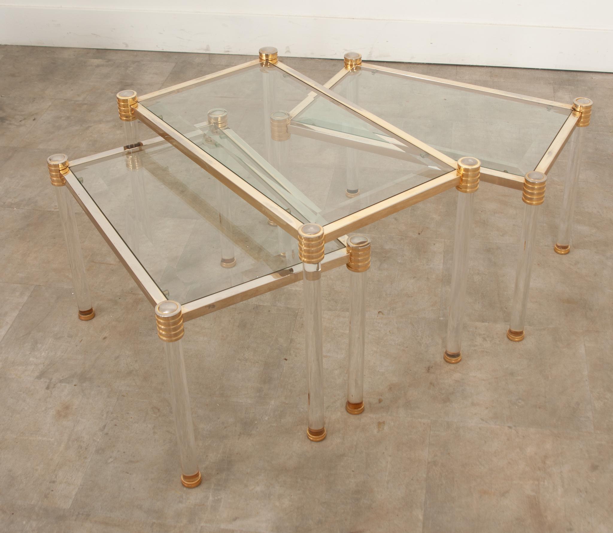 A fabulous vintage set of three French brass and glass side tables with unique flair. Their frames are made of patinated brass supported by lucite cylindrical legs with decorative brass caps. Snuggly enclosed in brass, the beveled glass tops are in