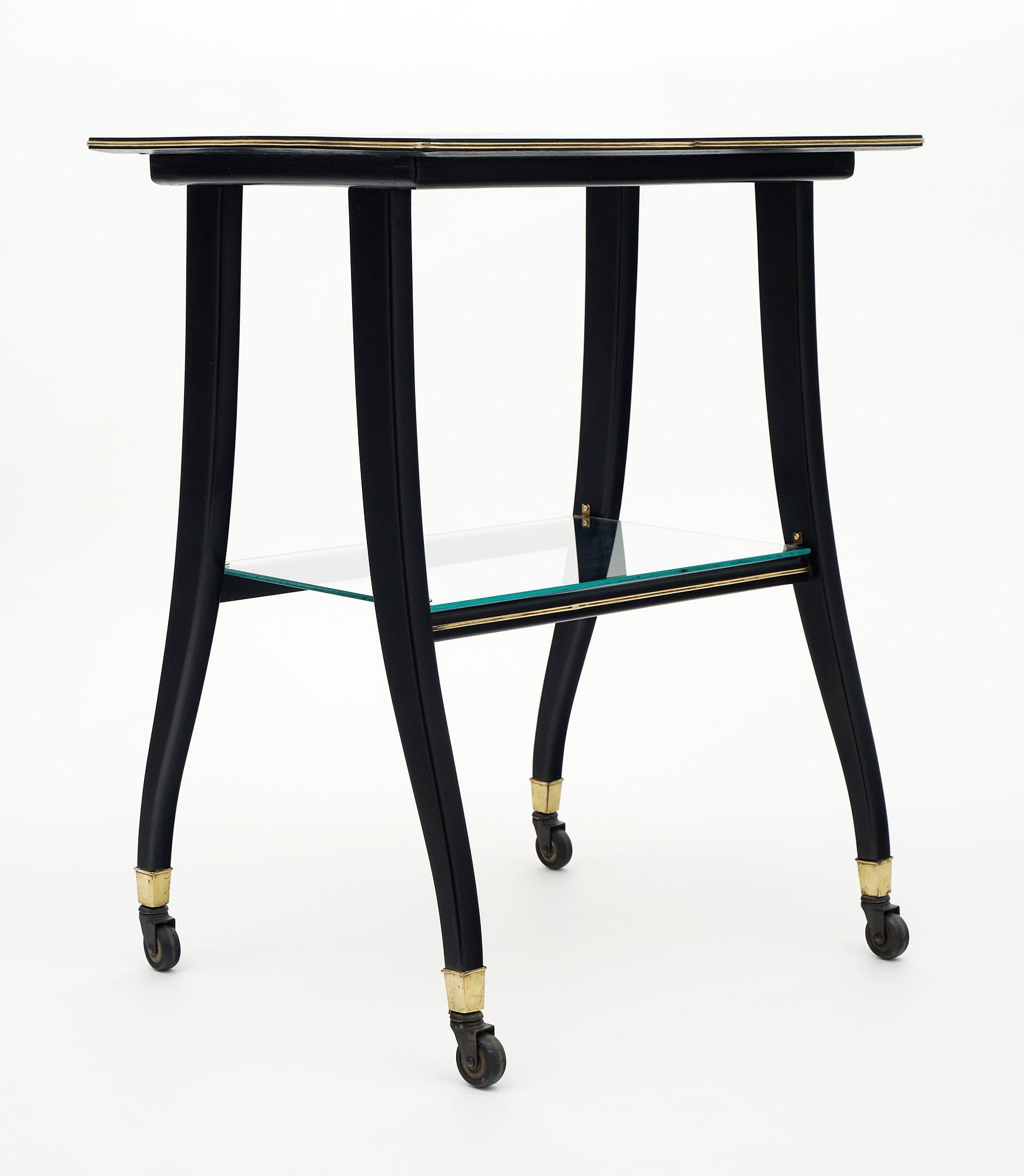 Side table on wheels made of mahogany veneer. We love this French Modernist piece with cabriole legs; brass feet; and brass trim around the top. The bottom shelf is glass. It is finished with an ebonized French polish.