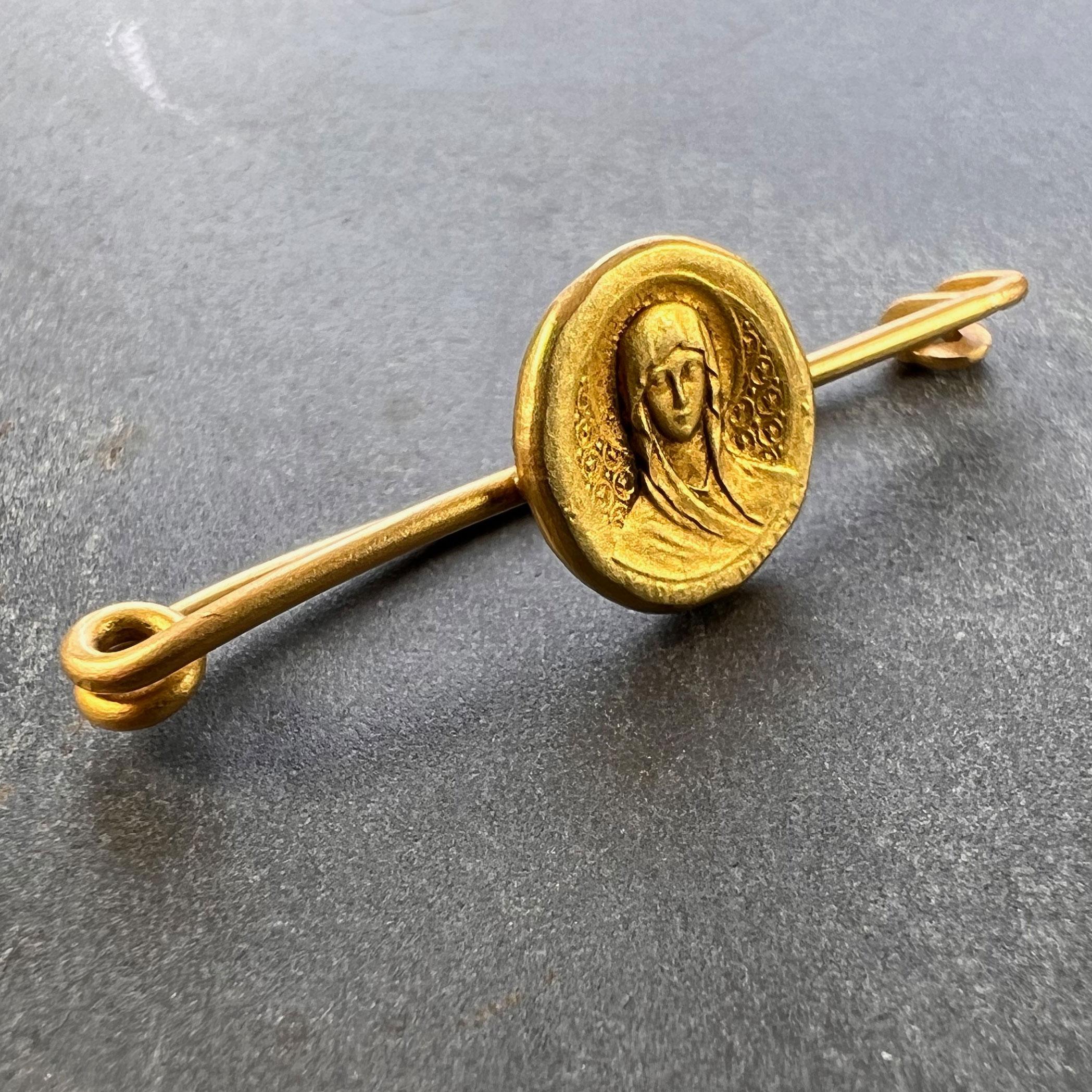 An 18 karat (18K) yellow gold pin brooch of a medal depicting the Virgin Mary with a halo on a repeating geometric background of encircled fleur de lis, with the phrase 'Virgo Maria' below. 

The reverse depicts the Marian symbol of the entwined