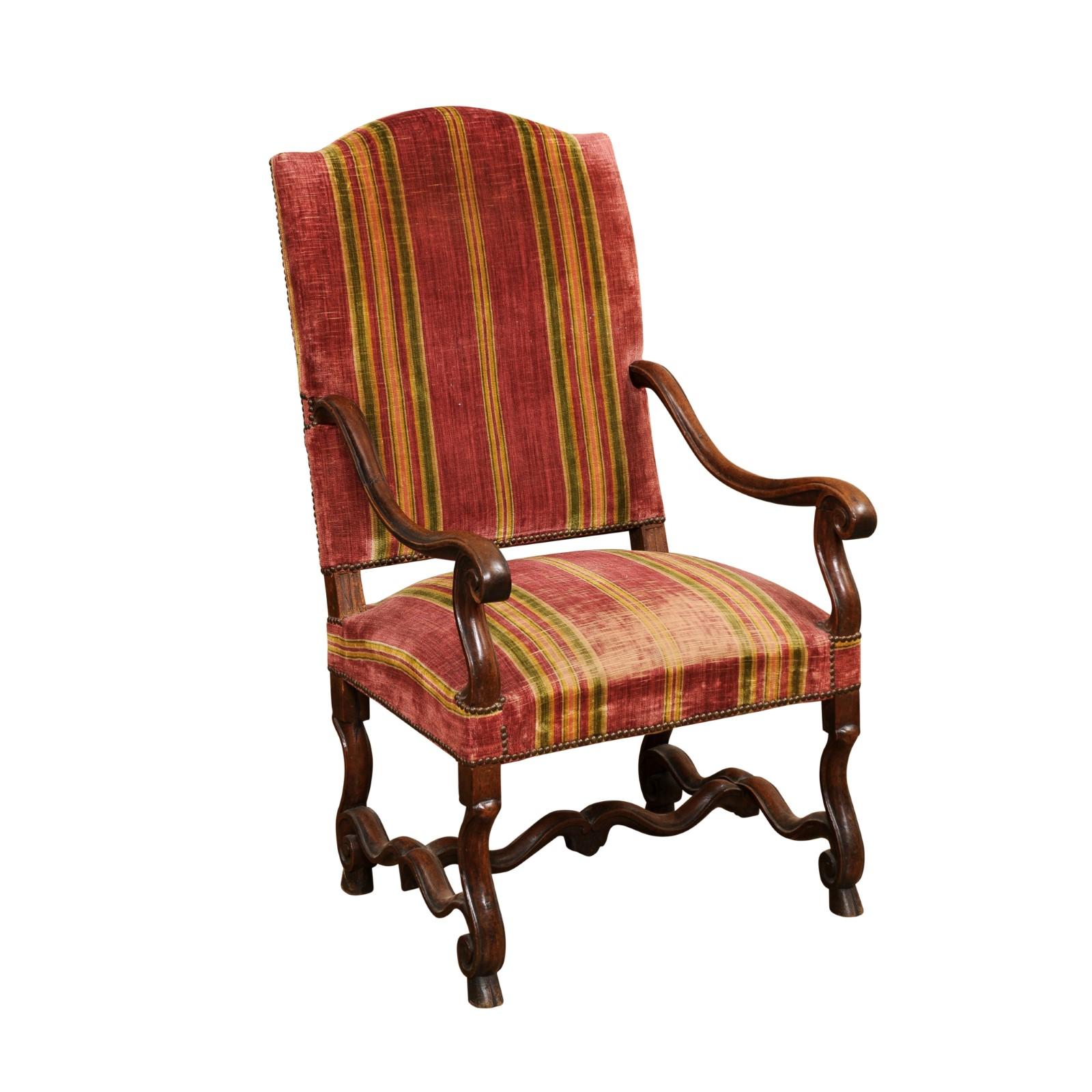 A French Louis XIV style walnut fauteuil circa 1790 with camelback, large scrolling open arms, Flemish scroll legs, curving H-form stretcher and old upholstery. Presenting a French Louis XIV style walnut fauteuil, circa 1790, a piece that exudes