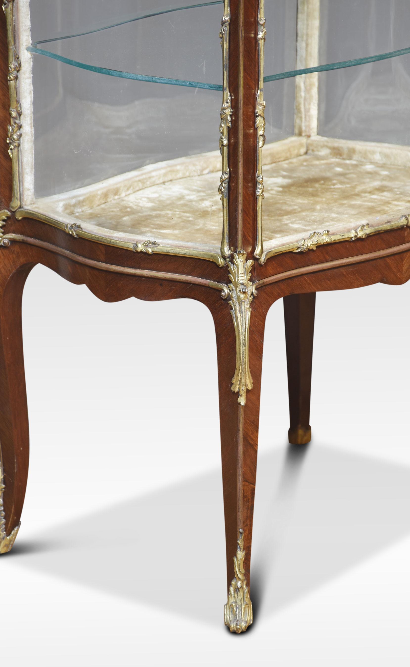 Meuble de bijouterie en noyer de style Louis XVI, de forme serpentine ornée de montures en métal doré. Le plateau en verre biseauté et les côtés vitrés façonnés mènent à la grande ouverture à une porte qui révèle deux étagères en verre. Le tout