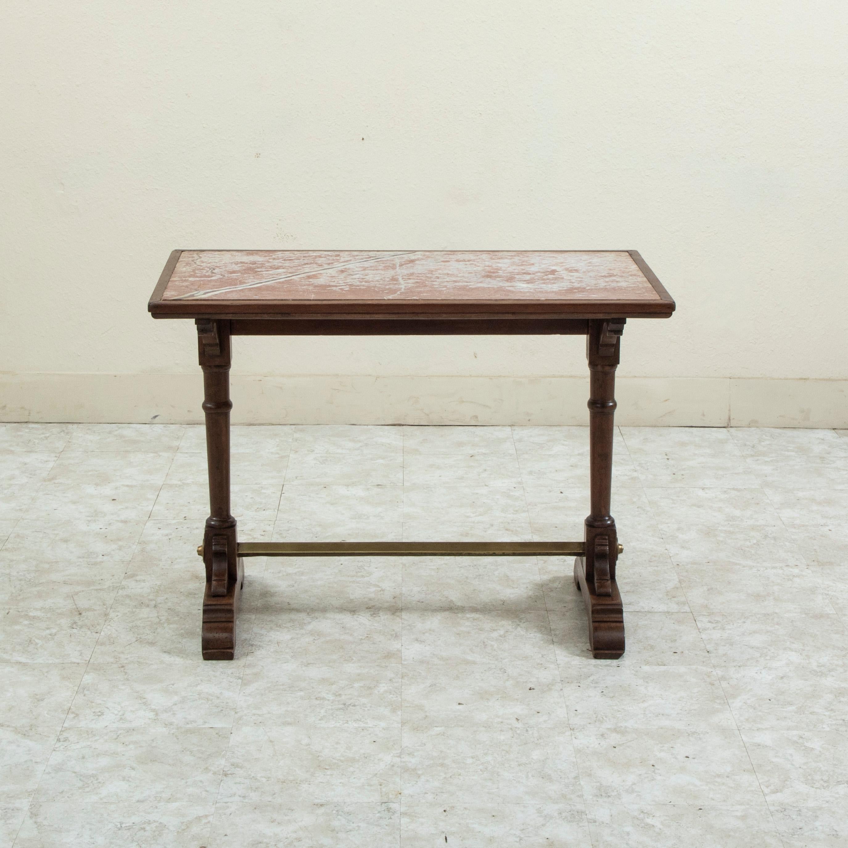 This French walnut bistro table or cafe table from the turn of the twentieth century features a rose colored marble top with grey and white veining. The top is surrounded by wooden trim and rests on turned legs. A brass stretcher connects the two