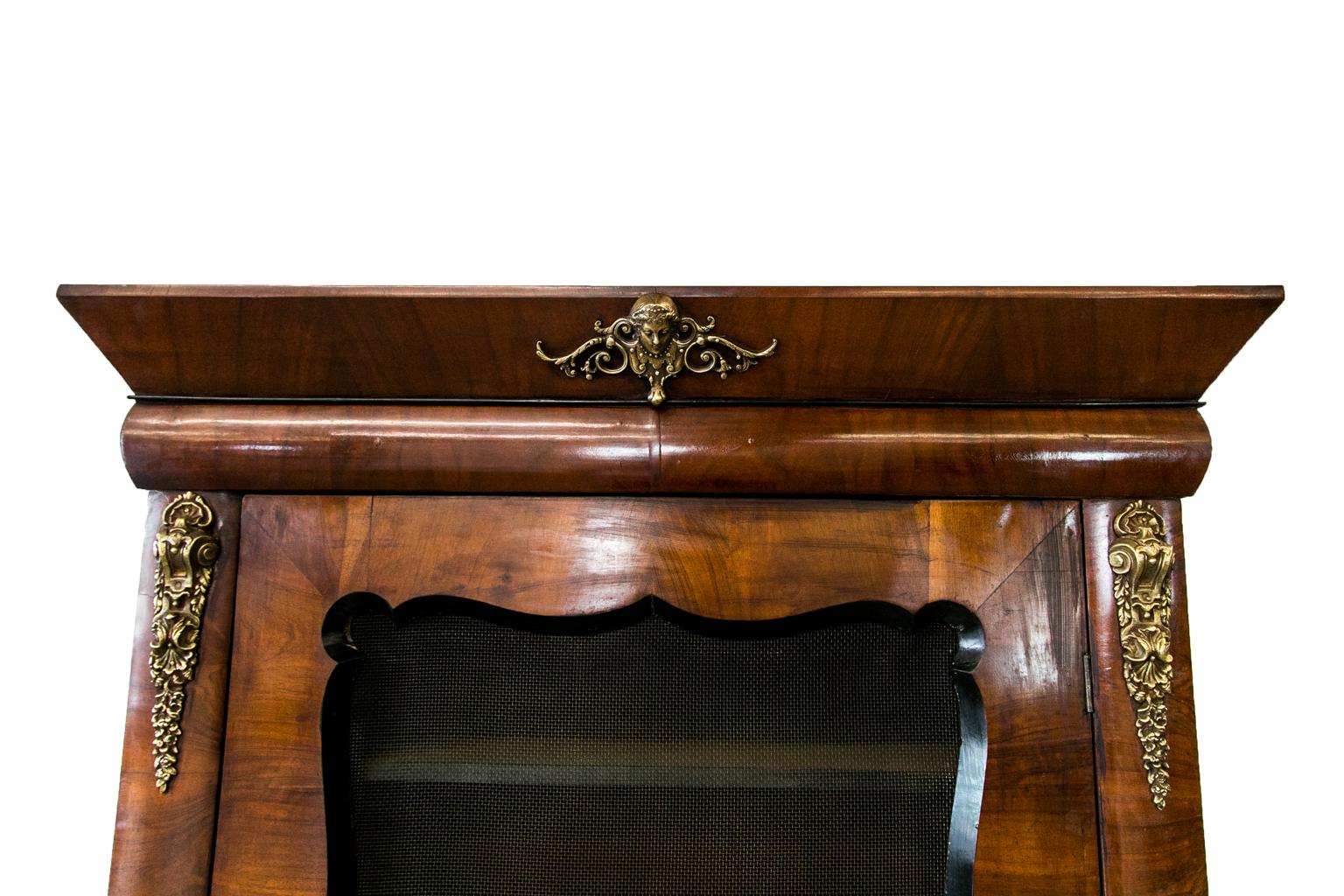 This French walnut bookcase has a wire mesh door with beveled ebonized scalloped framing. The stiles and frieze have convex banding in heavy cast brass ornamentation, four interior shelves. The stiles terminate in ebonized scroll feet. It has the
