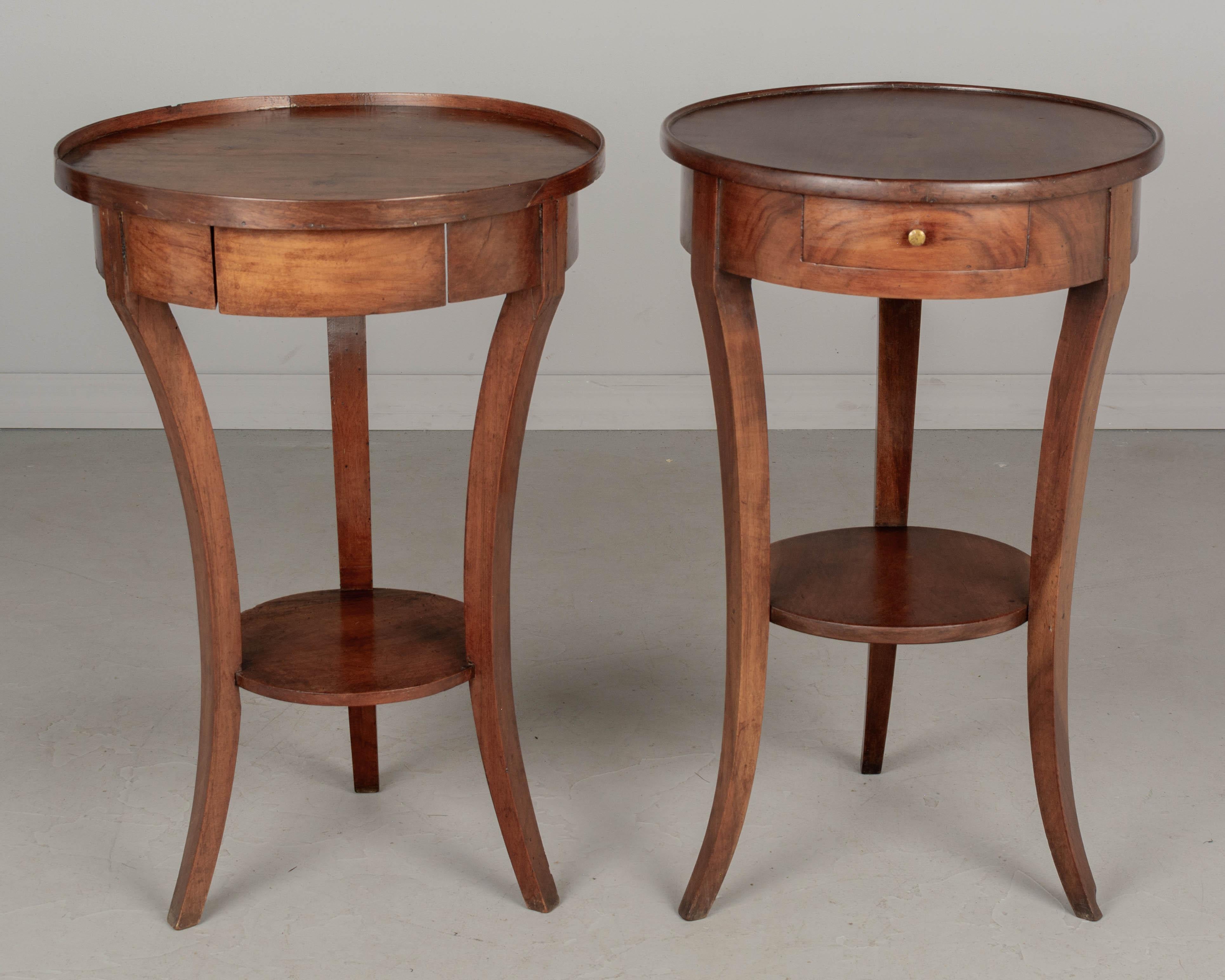 A set of two similar French walnut circular side tables, similar in scale, each with a dovetailed drawer, tripod base and lower shelf. One from the late 19th century and the other from the early 20th century. Each in good condition, the older one