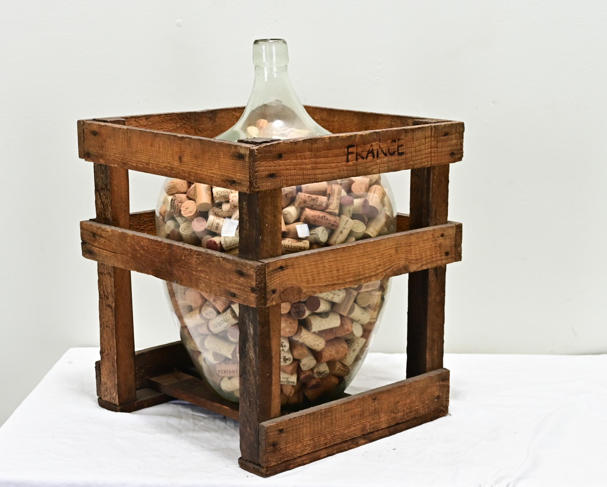 Questo barilotto per vino, o damigiana, in vetro trasparente e soffiato a mano del XIX secolo contiene una collezione di tappi di sughero. Su una cassa di quercia fatta a mano è impressa la scritta 