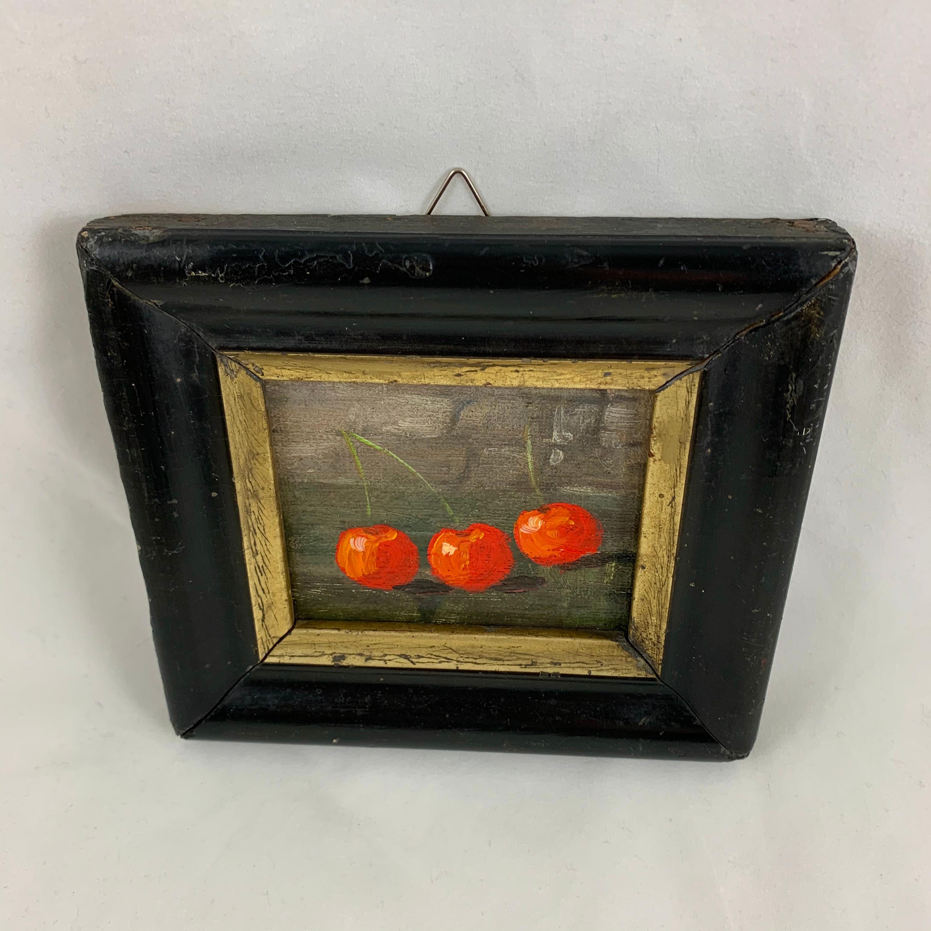 Found in France, a charming, early 20th century French still life of three red cherries, Les Trois Cerises, painted in oil on board. The cherries pop brightly from the background that faintly shows a stone wall. Presented in a wide wood frame,