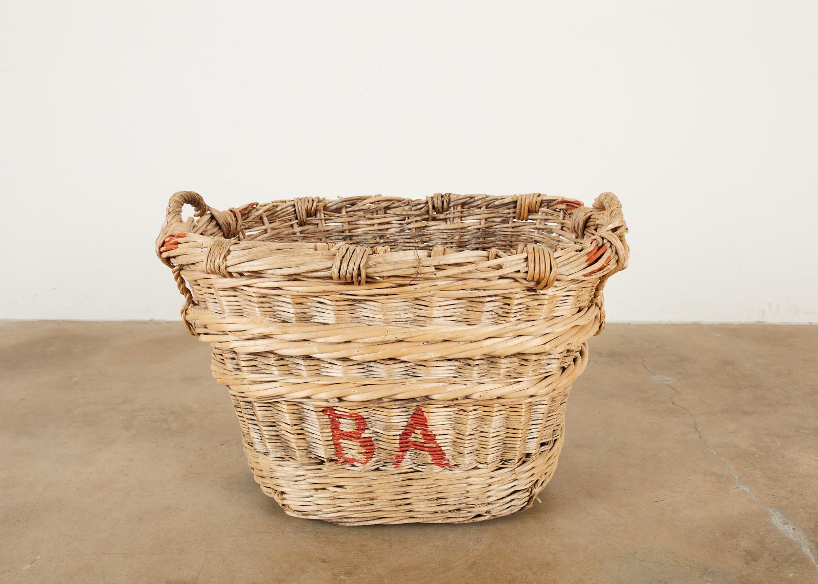 Rustic French chateau champagne grape harvesting basket constructed from woven wicker reeds. Features a thick braided top with handles on each end and brightly colored champagne house initials on the sides reading BA in vivid red paint. Reinforced