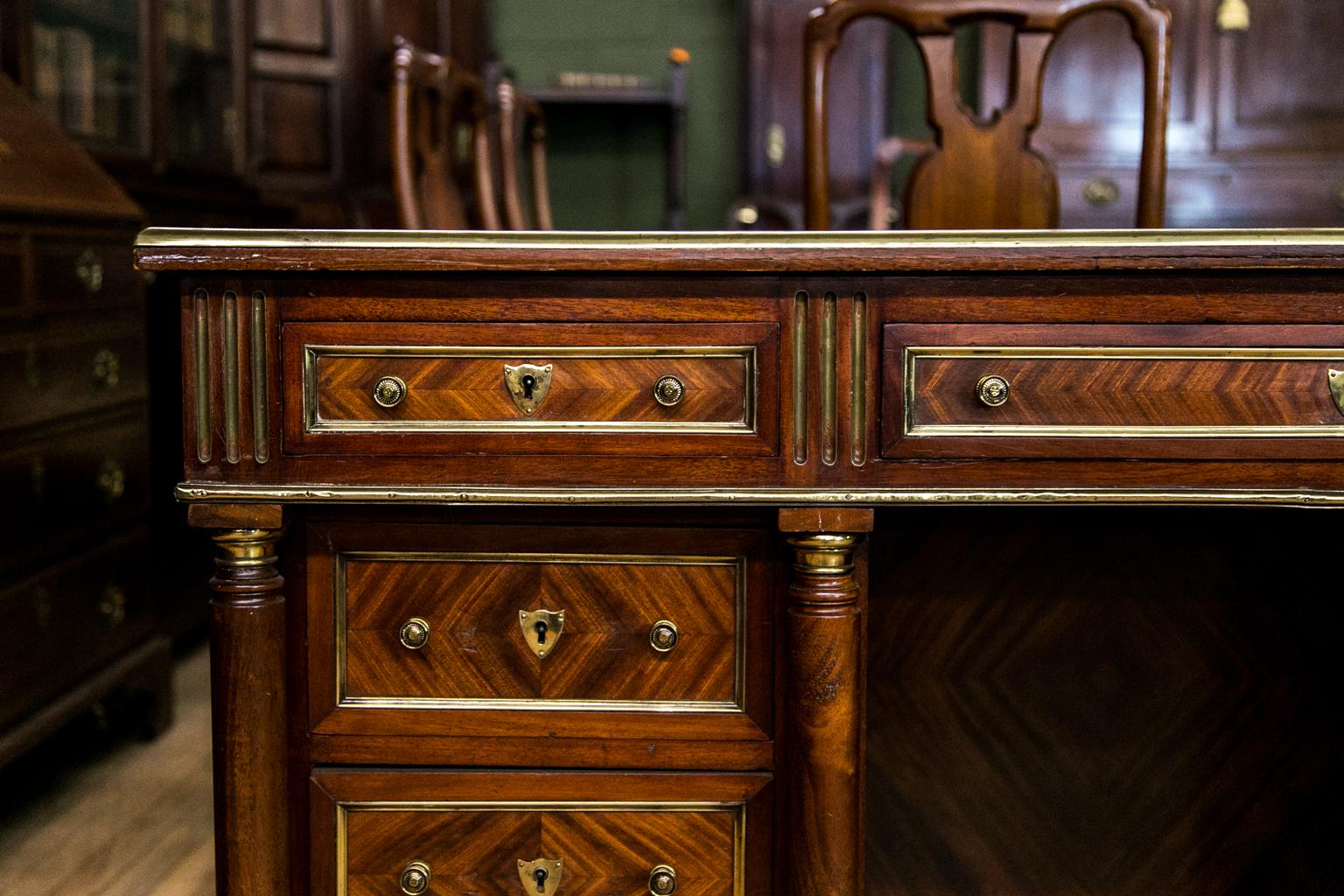 This French writing desk has a bookmatched mahogany top, drawer fronts, sides, back panels, and the interior of the knee hole. It is overlaid with brass moldings on all four sides. The top has a green, gold, and blind tooled leather top with pull