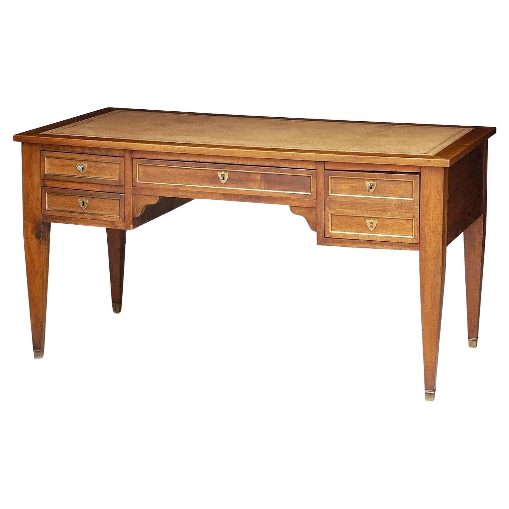 A fine French writing table or desk of walnut and mahogany, featuring an embossed leather top, four drawers with brass escutcheons, and resting on tapering legs with brass caps.
