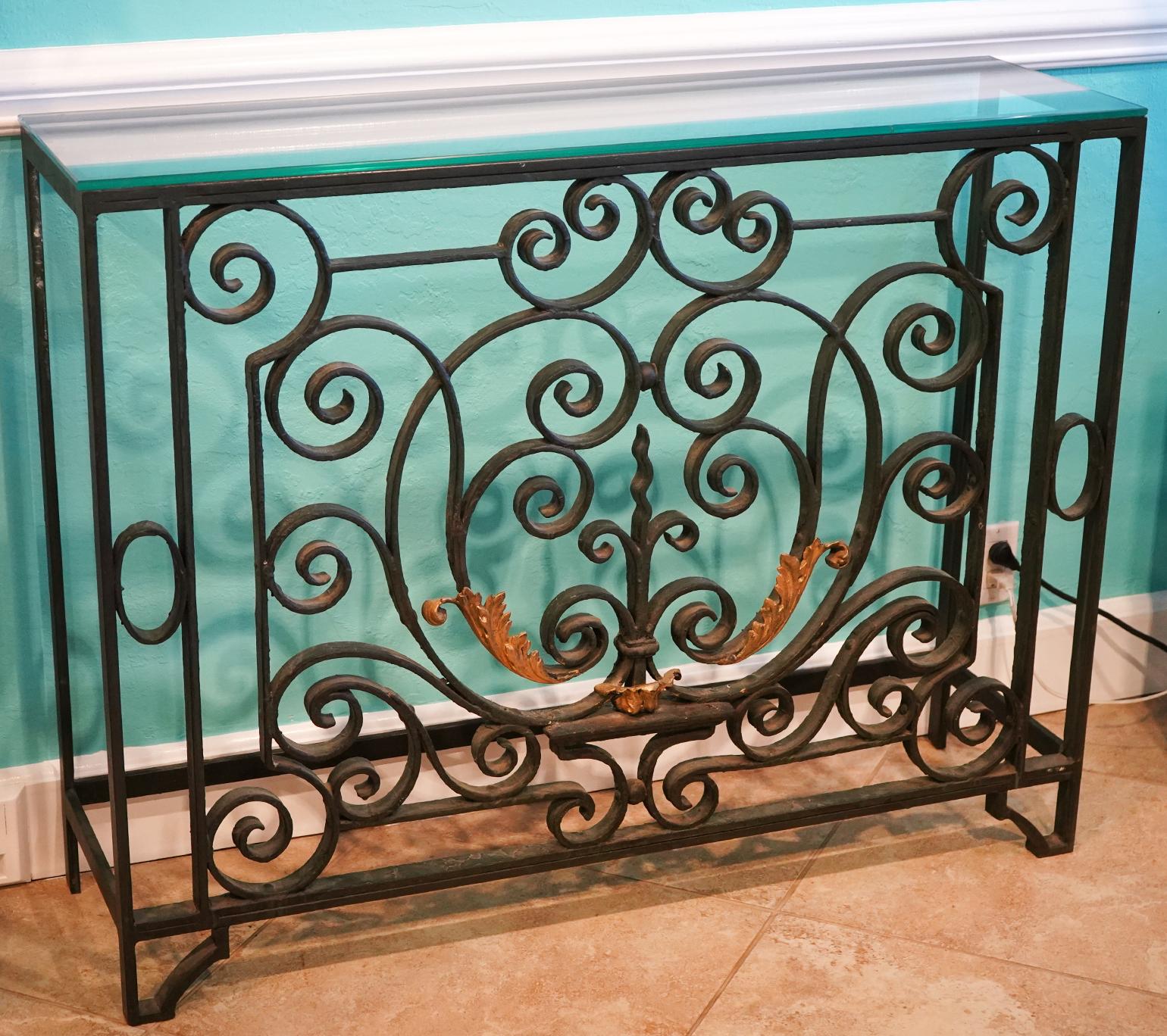 This elegant wrought console table in the French tradition features a front with unfolding scrolls centering a stylized fleur de lis adorned by gilt leaf work. The latter being of great decorative effect. The top is made of tempered glass and is