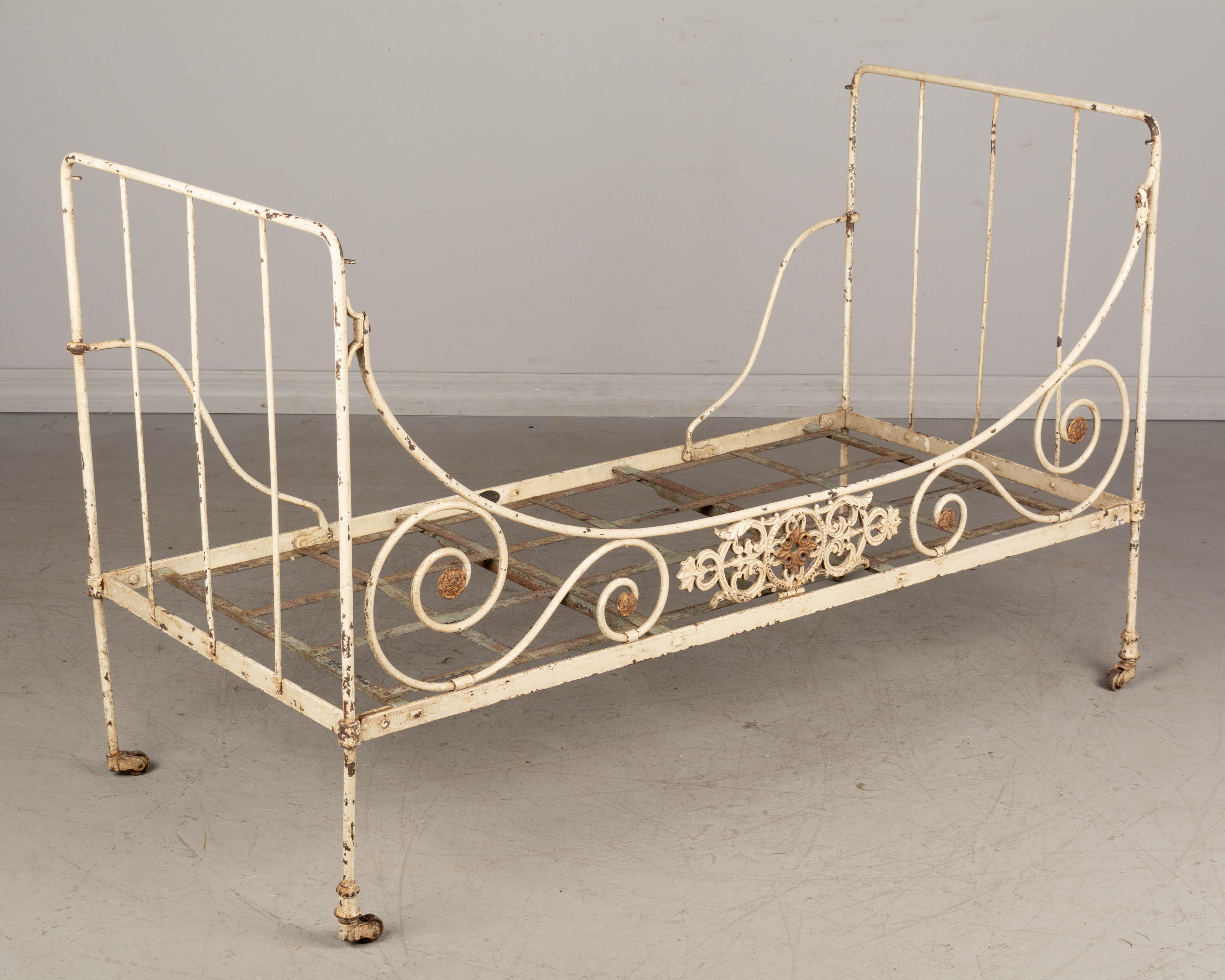 A French wrought iron child's bed or small daybed with white painted patina. Cast iron decorative element with gold painted medallion. Woven iron straps support the mattress (not included). Oval metal tag: F. Brevette. Some paint loss and rust.