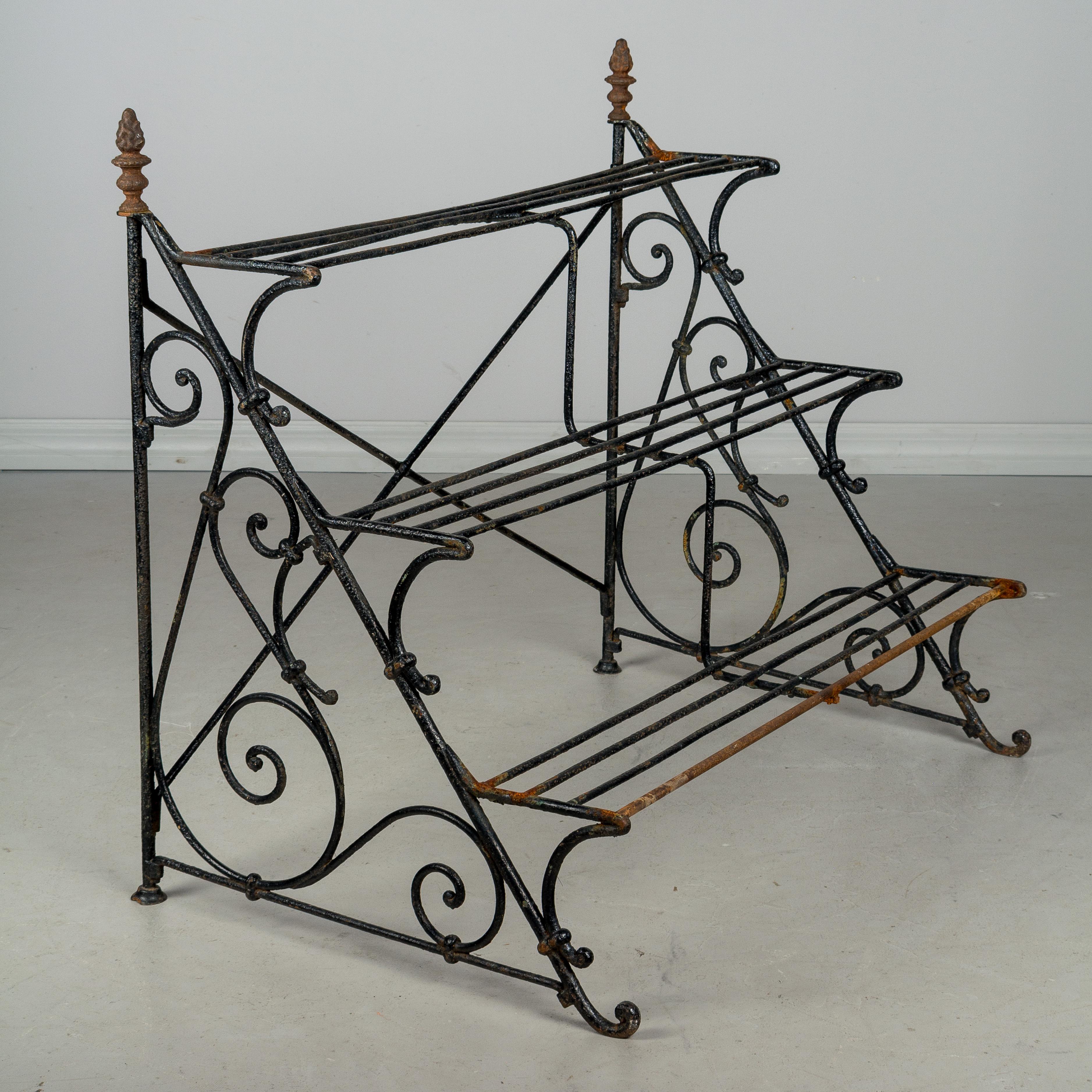 A French wrought iron stepped garden shelf for potted plants. Scroll design on the sides and decorative finials at the top. Old black painted patina is rusted in places especially on the lower step and the finials.