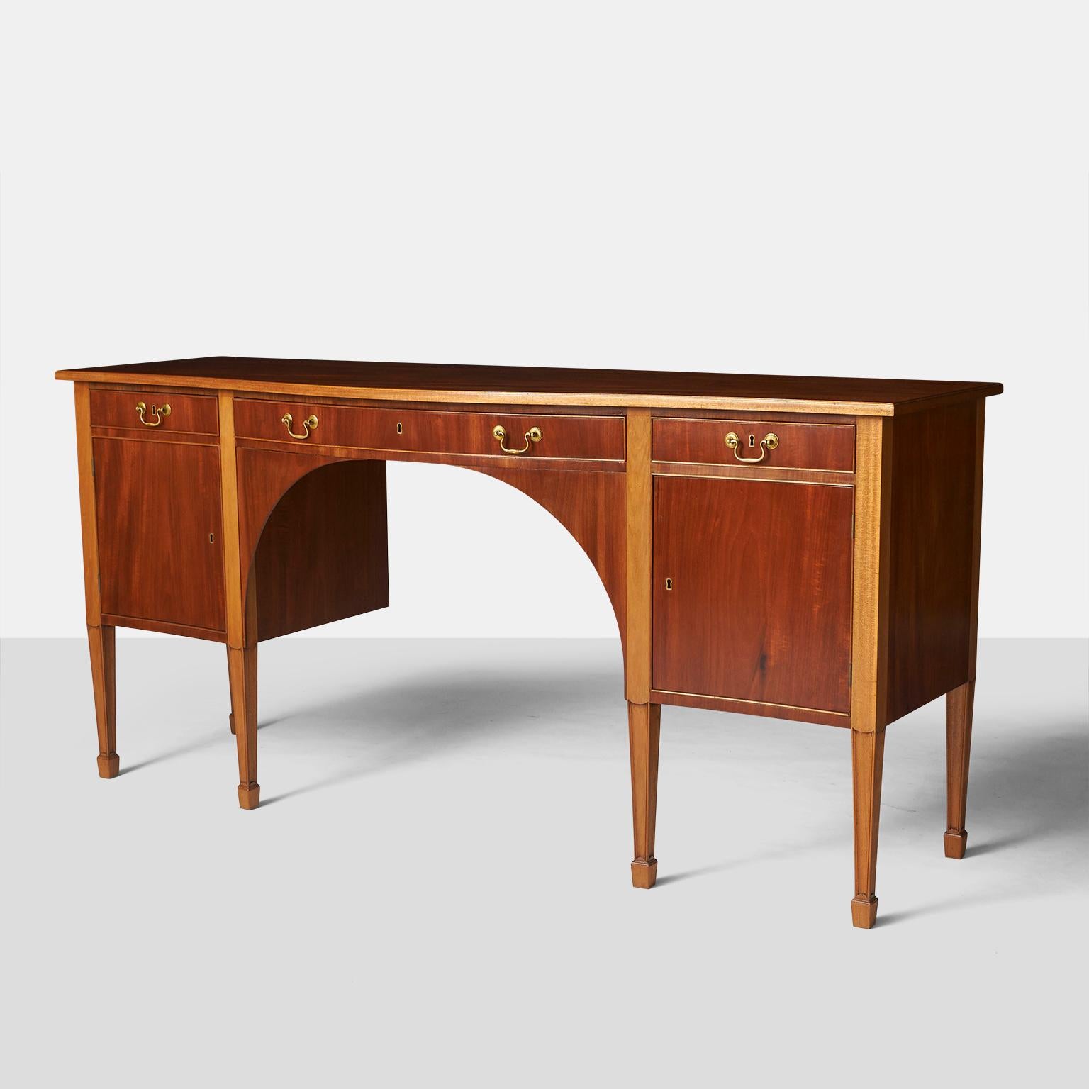 A mahogany Sheraton style sideboard, lightly curved front with three drawers and two doors behind which there are shelves. Tall tapered legs. Brass pulls. Key included.