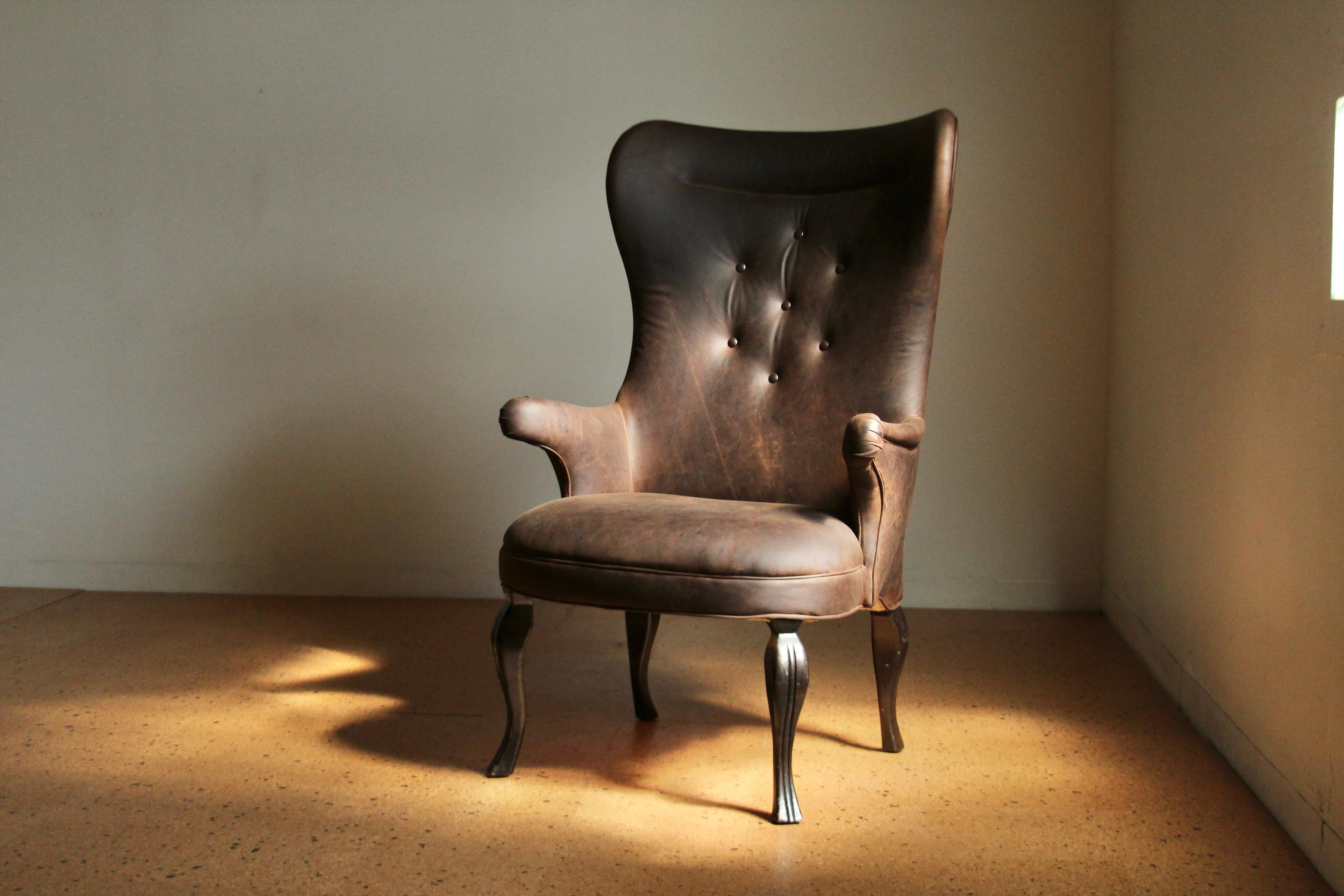 Un grand fauteuil à oreilles vintage à la manière de Frits Henningsen, avec un haut dossier incurvé, des accoudoirs pointus, des capitons et de hauts pieds incurvés avec des détails sculptés. Cette pièce unique et de grande taille a été refaite en