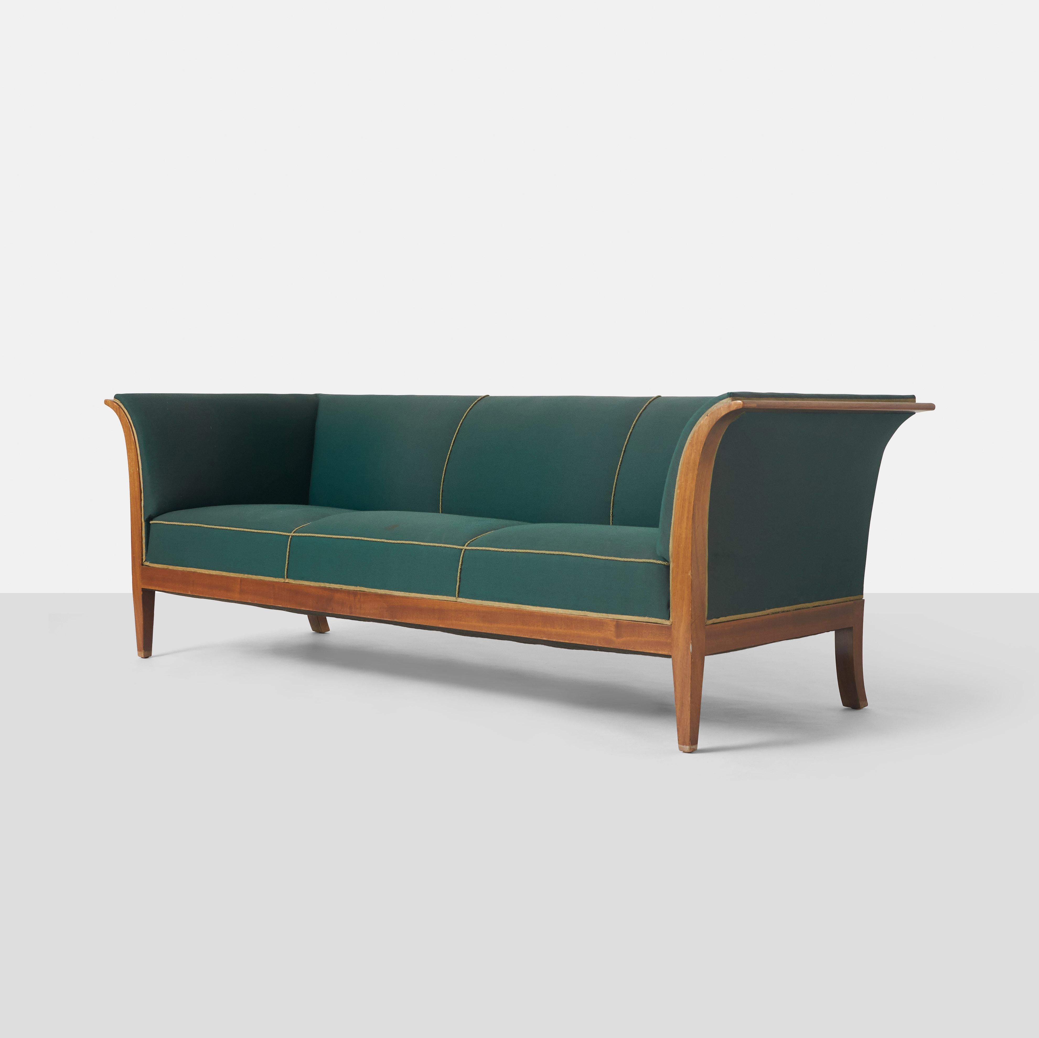 A Frits Henningsen three seater sofa on a mahogany frame with eight legs and covered with green wool. The sides have exposed wood panels on either side. It was designed and made circa 1938 by Danish cabinetmaker Frits Henningsen

