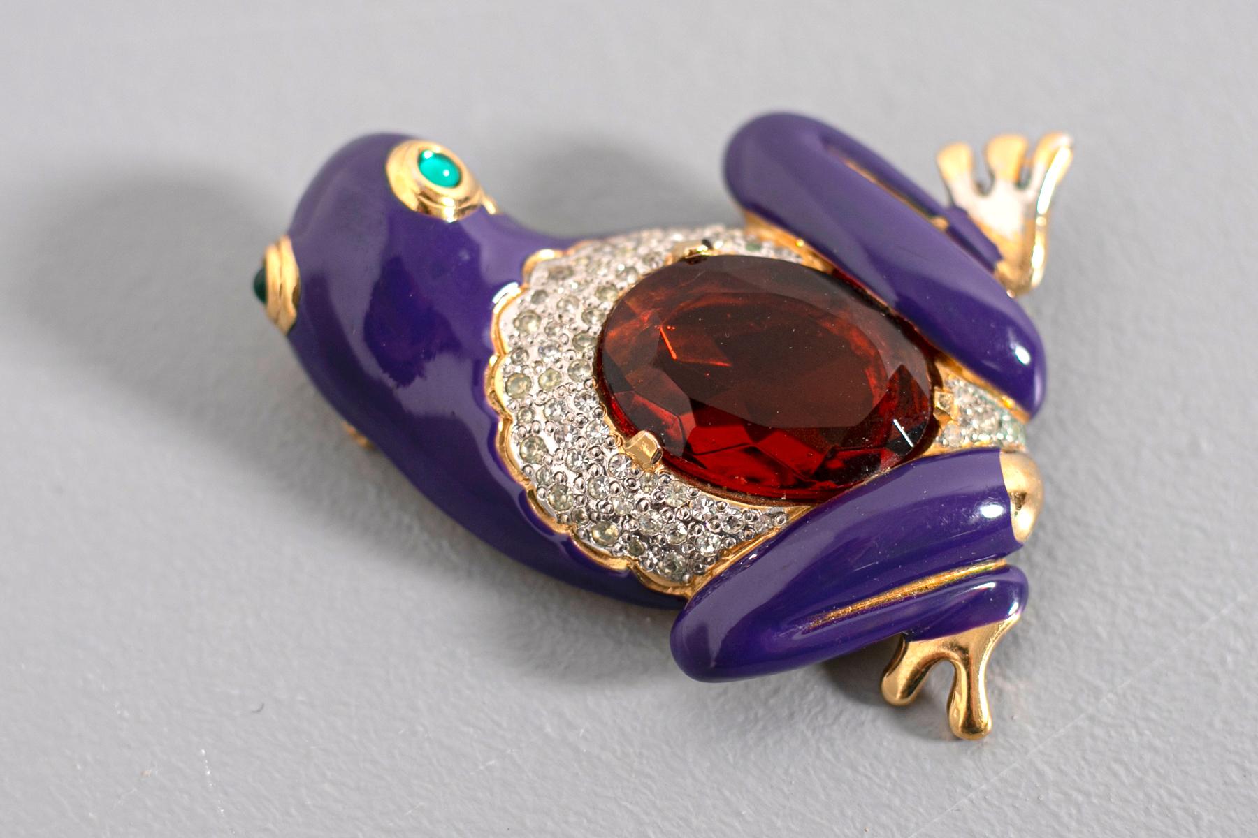 Magnifique broche bijou signée par le grand designer Valentino des années 80.
En forme de grenouille, sur une base en métal de couleur or et émaillée violet, au dos est agrémenté d'une grosse pierre rouge et de petits strass argentés, les yeux de la