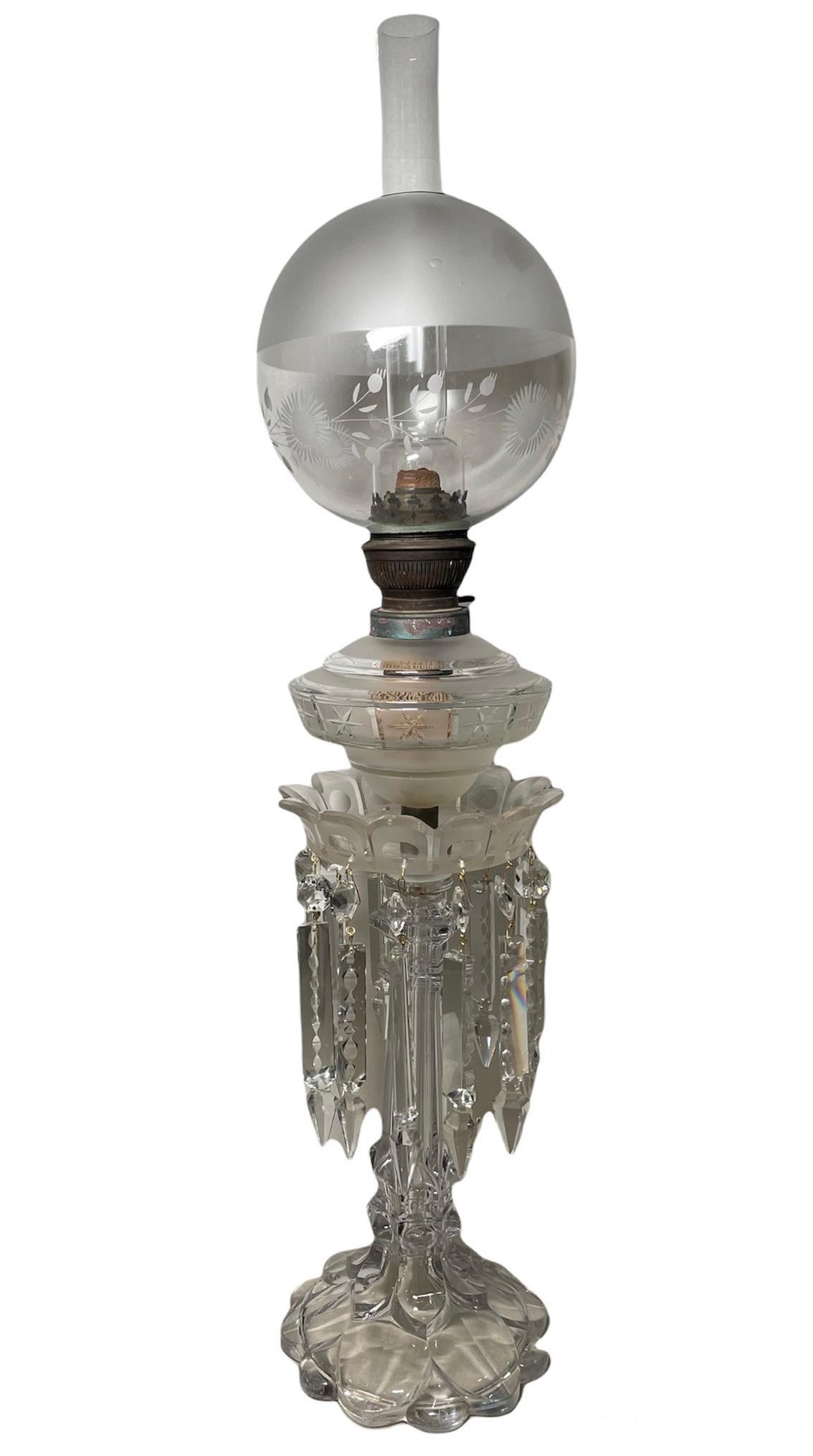 This a clear/frosted cut crystal banquet oil lamp. It consists of a crystal lamp chimney, etched with flowers branches crystal ball shade joined to a brass burner with frosted crystal font cap decorated with a band of stars. This last piece has a